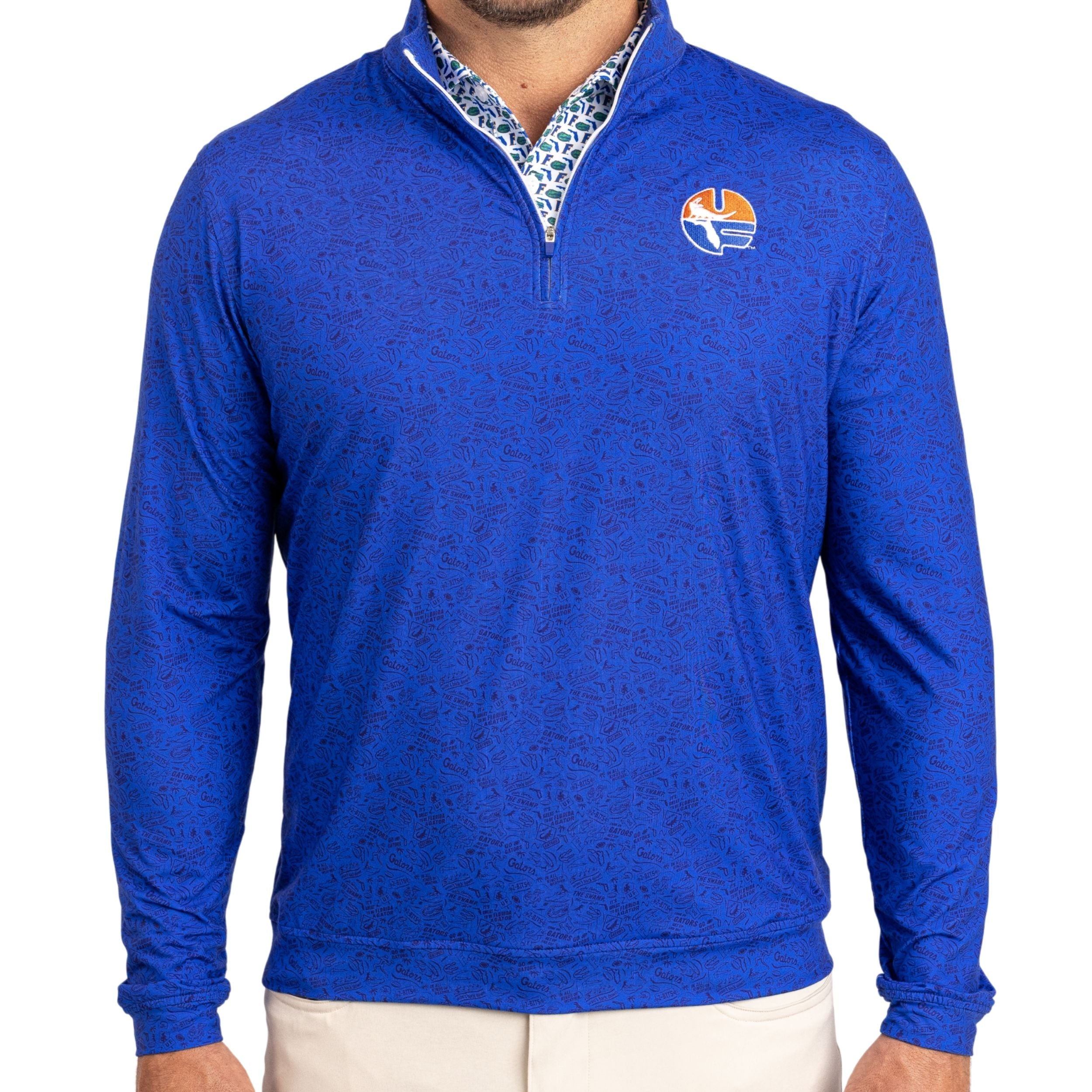 A man wearing a royal blue long-sleeve quarter-zip pullover with a subtle tonal pattern, featuring a Florida Gators logo on the left chest and a patterned inner collar, paired with light beige pants against a white background.