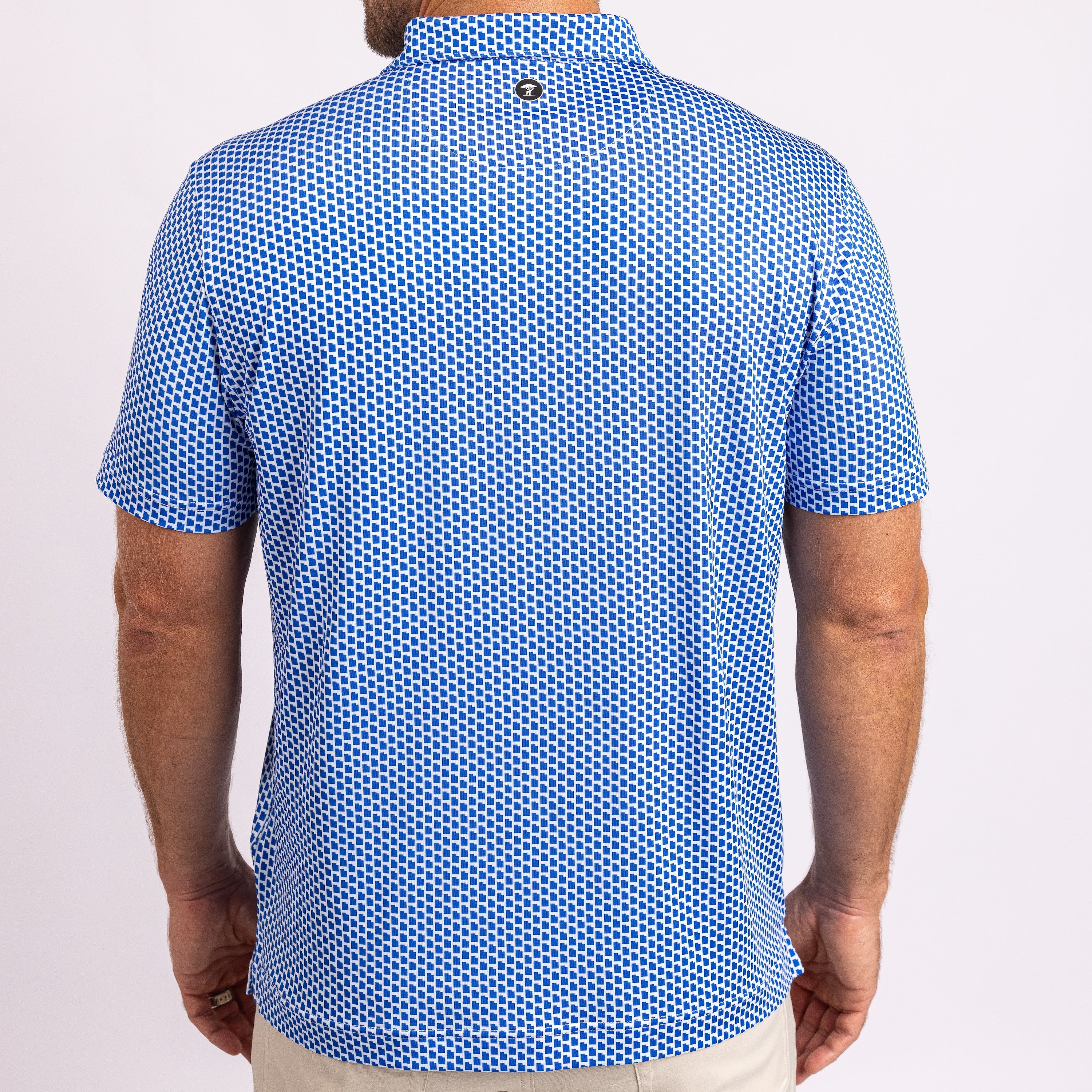A man viewed from the back wearing a short-sleeve polo shirt with a blue and white geometric pattern, paired with light beige pants, against a plain light background.