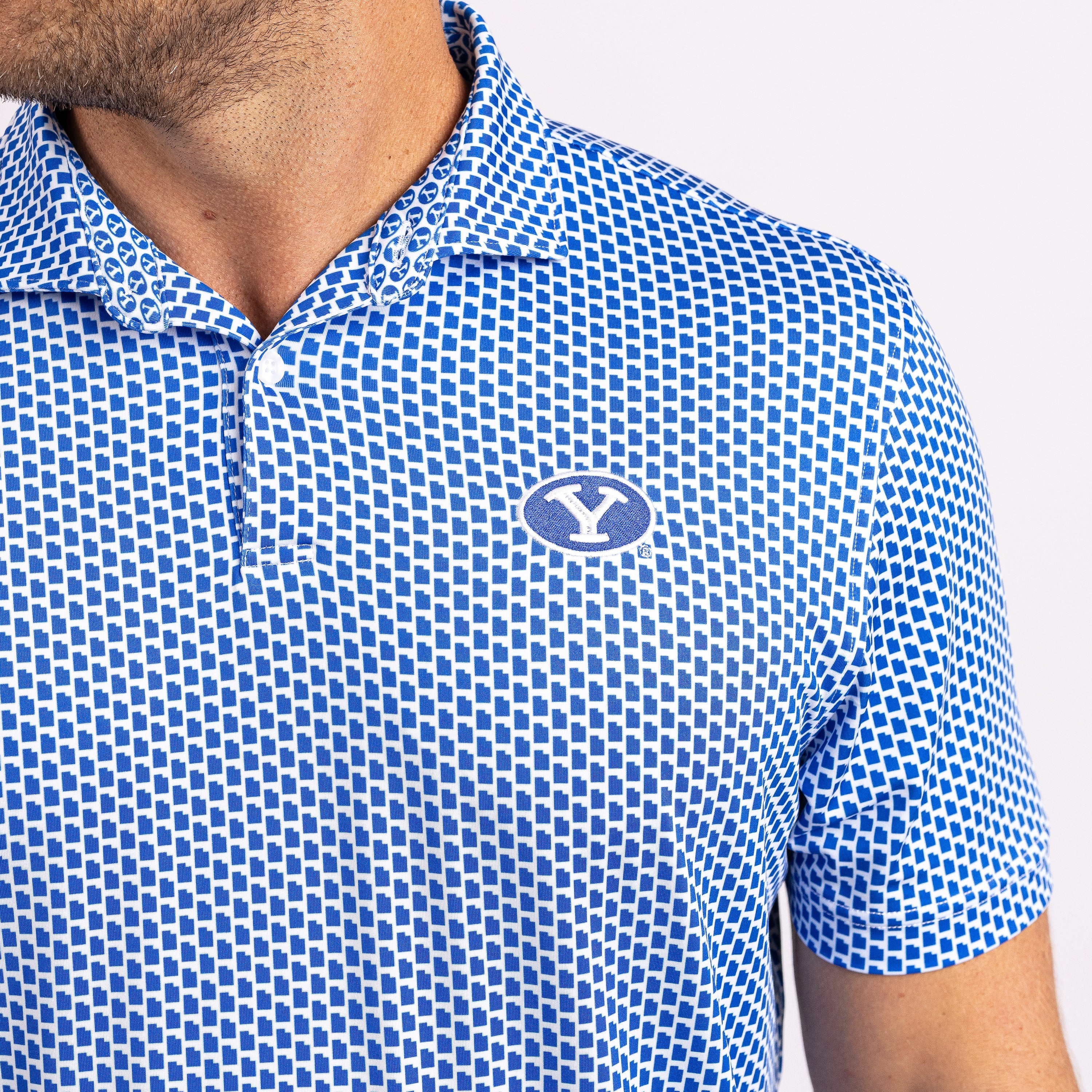 Close-up of a man wearing a blue and white patterned polo shirt with a blue and white logo on the chest, against a light gray background.
