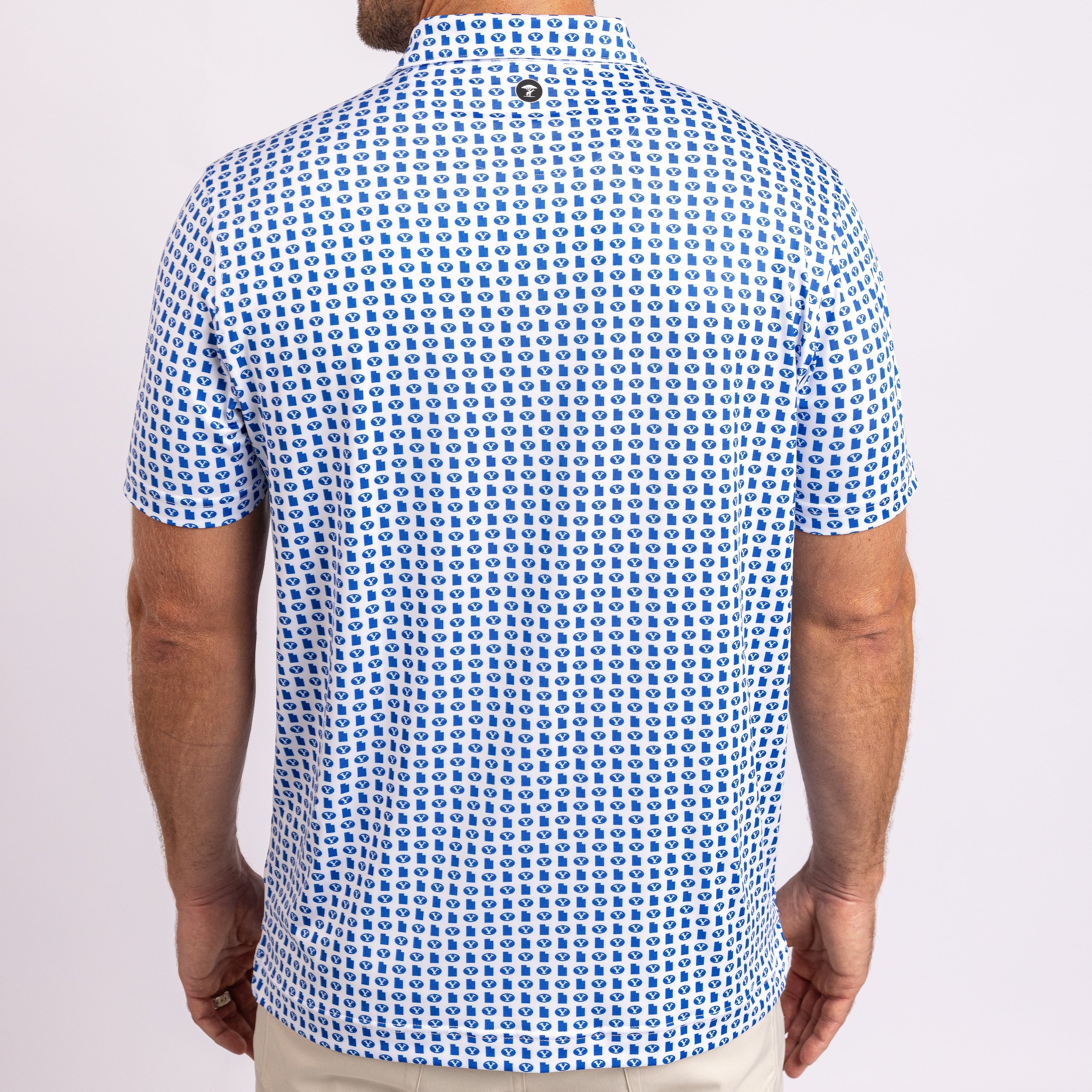 A man viewed from the back wearing a short-sleeve polo shirt with a dense blue geometric pattern on a white base, paired with light beige pants against a light gray studio background.