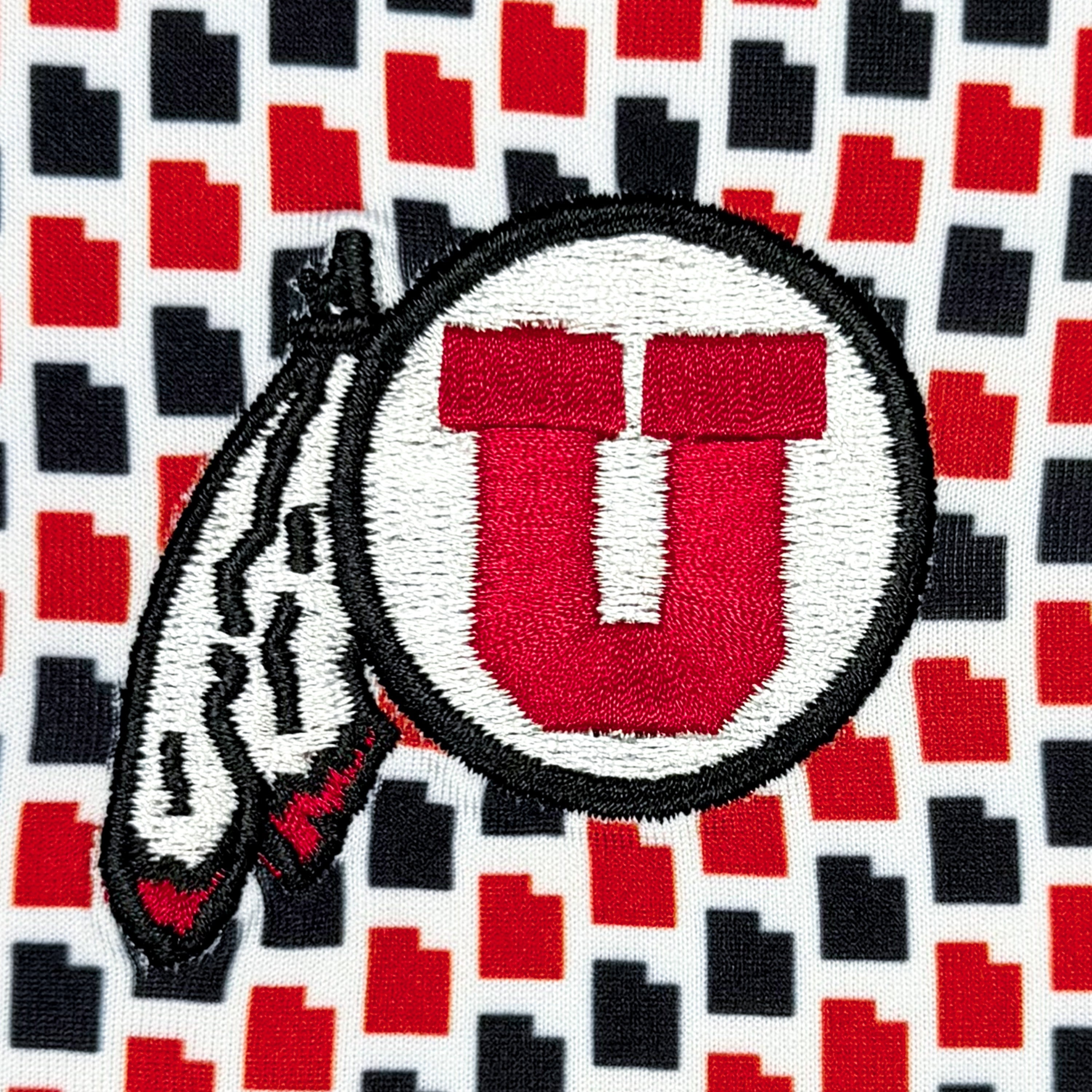Close-up of an embroidered patch featuring a red letter U inside a white circle with a black border, overlaid on a red, black, and white geometric patterned fabric.
