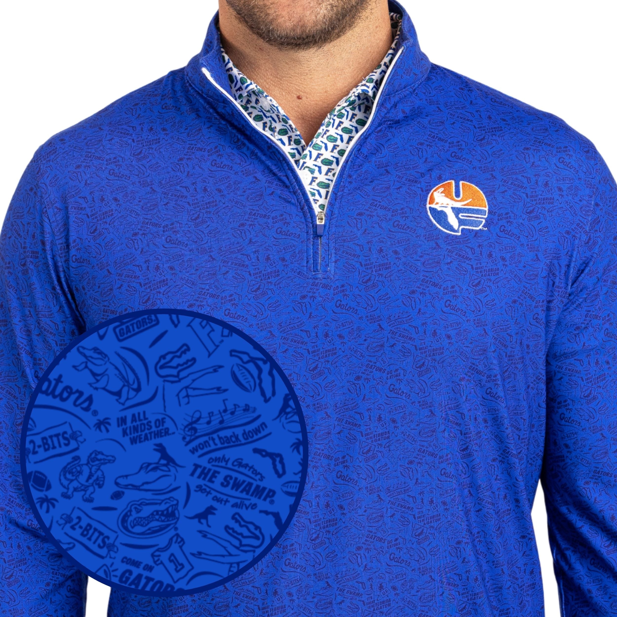 A close-up of a man wearing a royal blue quarter-zip pullover with an all-over subtle pattern. The shirt features a small circular logo on the left chest and a visible patterned collared shirt underneath.