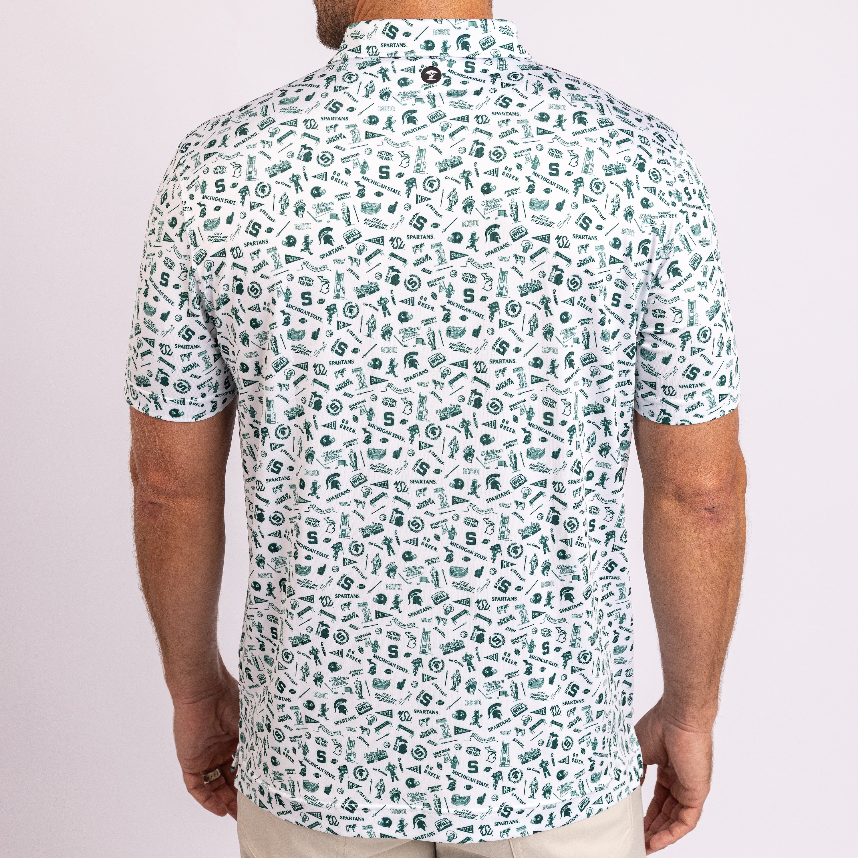 A man viewed from the back wearing a short-sleeve button-up shirt with an all-over green and white collegiate-style graphic print, paired with light beige pants against a light pink studio background.