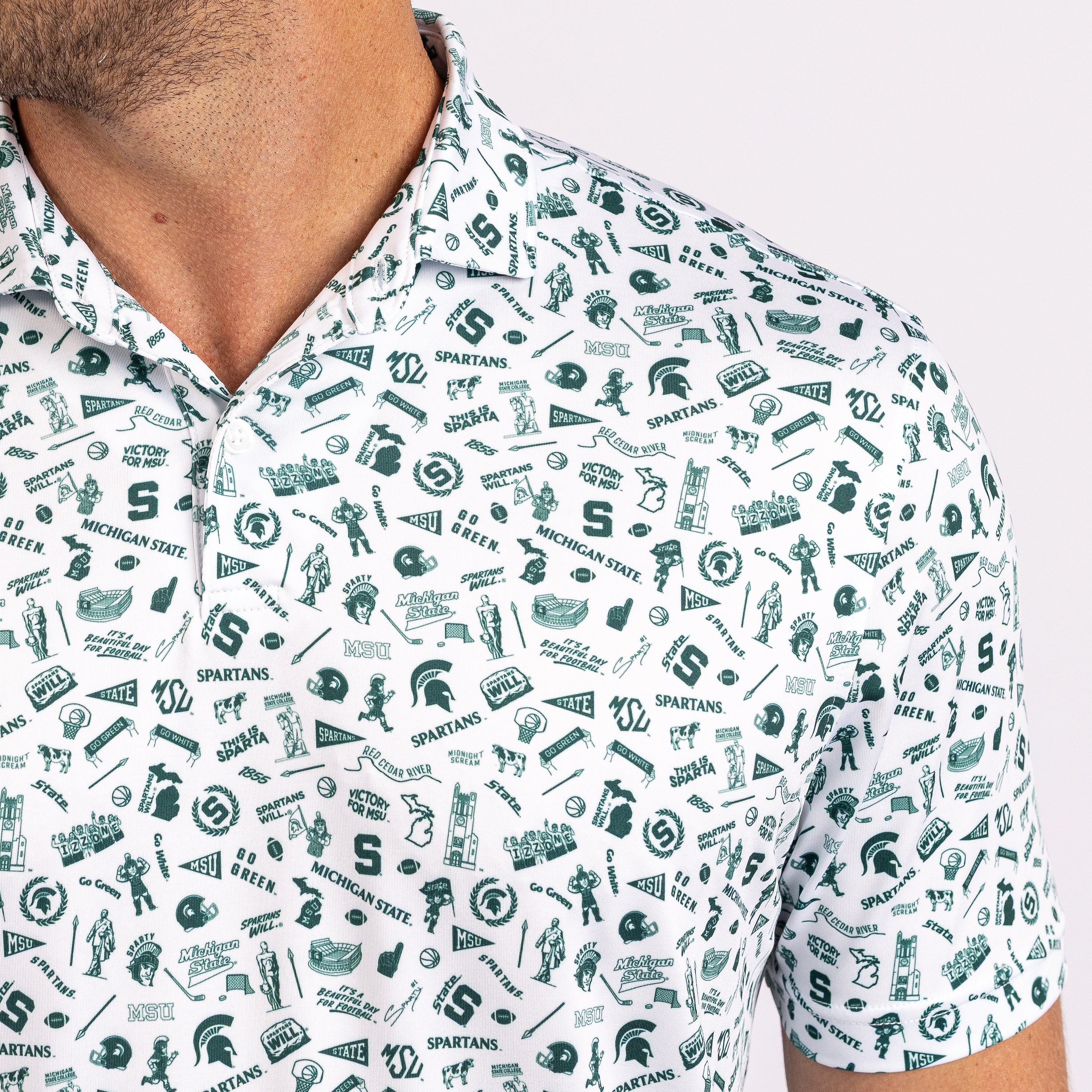 A close-up of a white short-sleeve polo shirt with an all-over green Michigan State Spartans-themed graphic print, worn by a model.