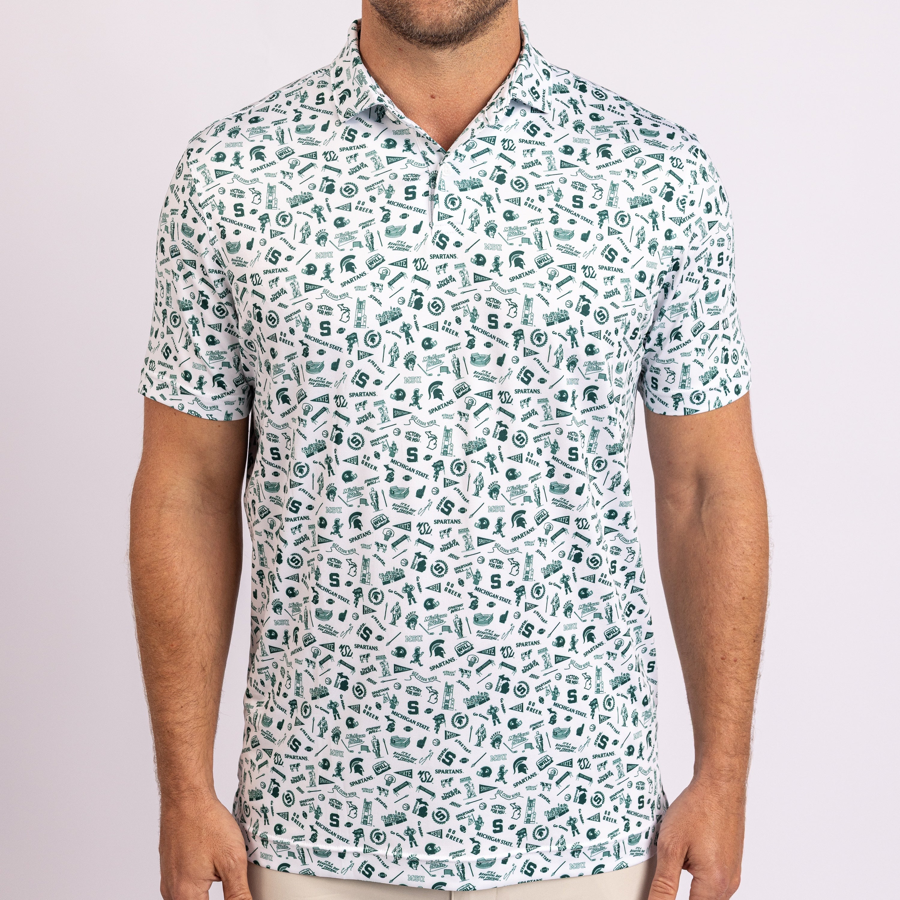 A man wearing a short-sleeve polo shirt with an all-over abstract pattern of green and black shapes on a white base, shown from the chin down against a light pink studio background.