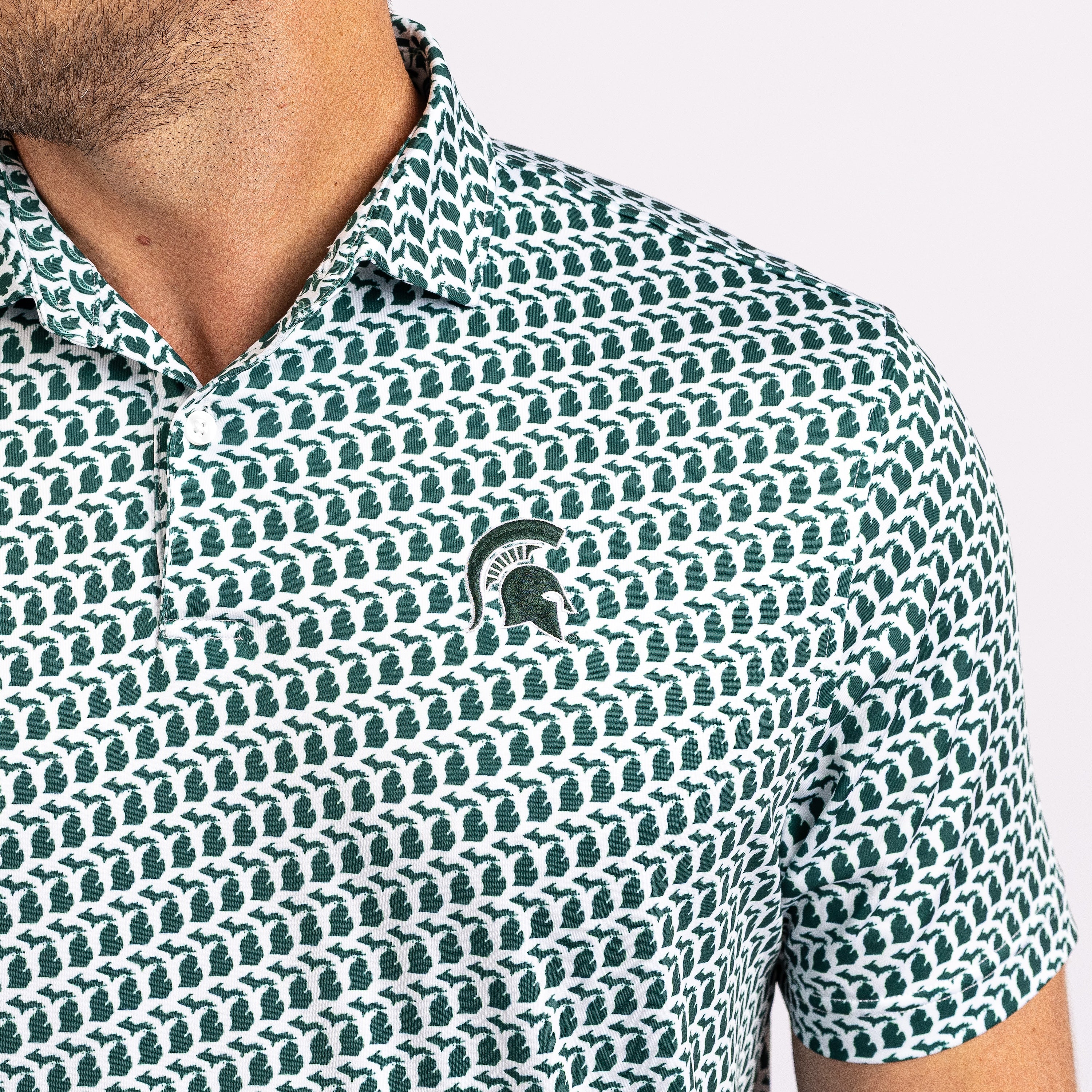 Close-up of a man wearing a short-sleeve polo shirt with a repeating green and white pattern and a small Michigan State Spartans helmet logo on the chest.