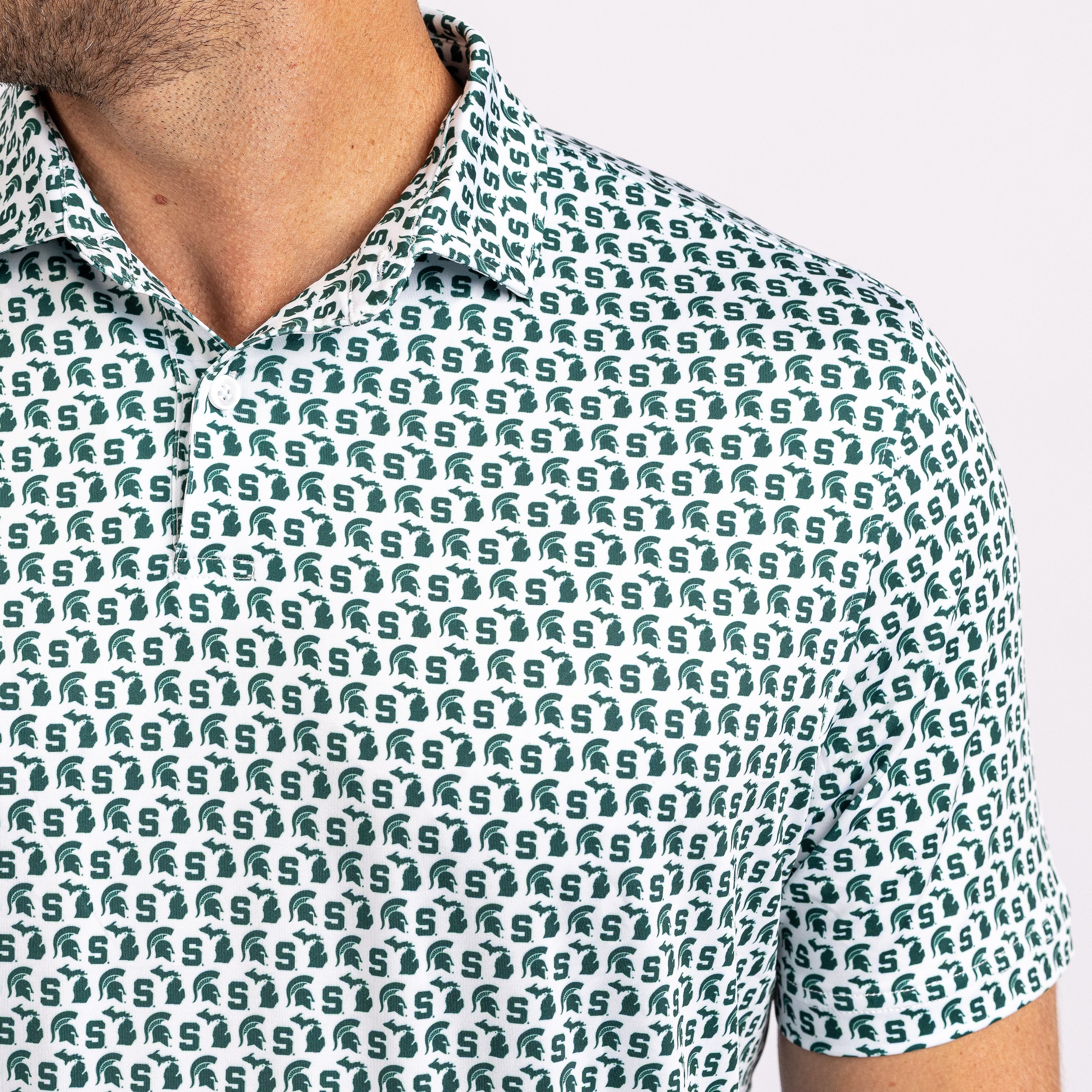 Close-up of a man wearing a short-sleeve polo shirt with an all-over repeating Michigan State Spartans logo pattern in dark green on a white base.