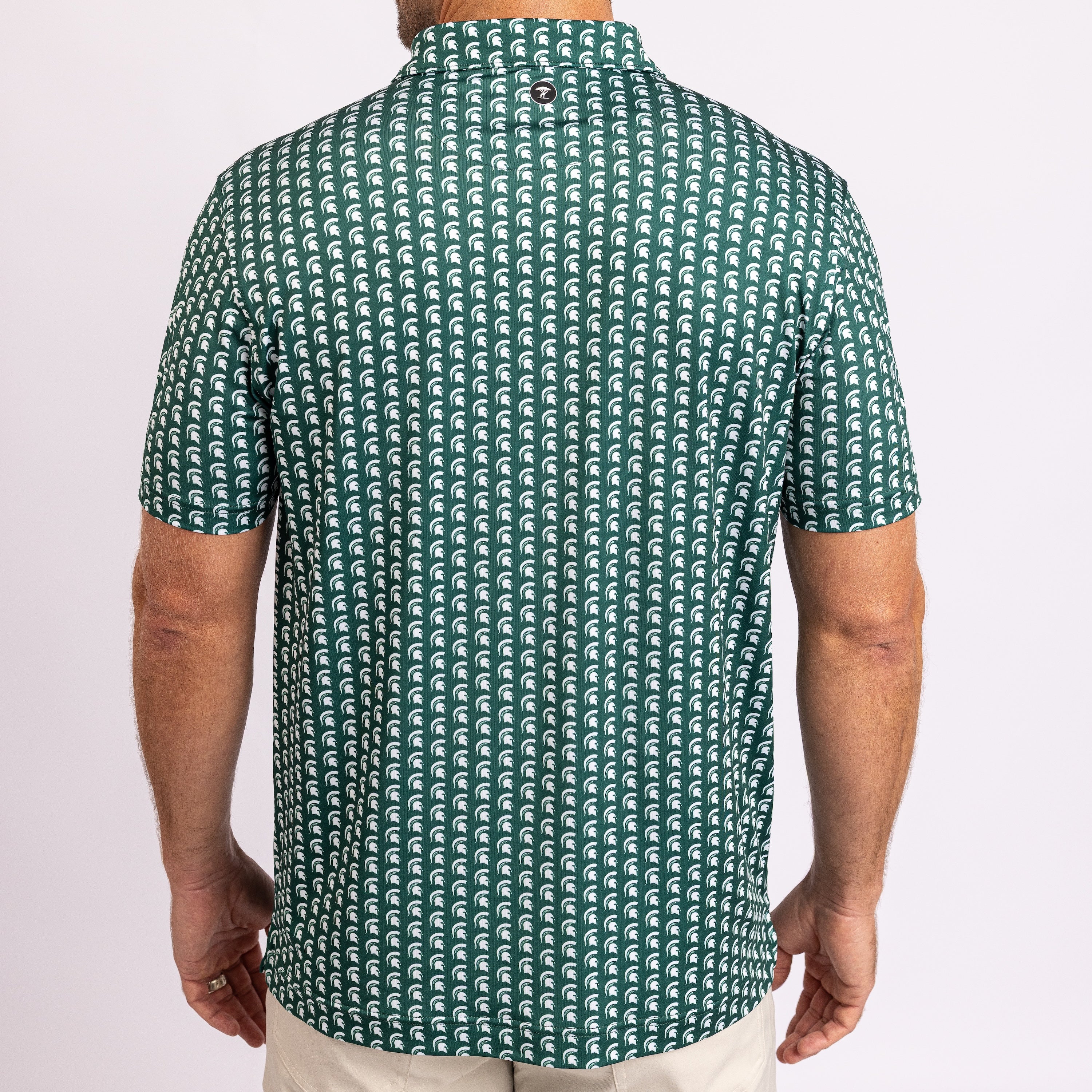A man viewed from the back wearing a short-sleeve collared shirt with an all-over repeating wave pattern in dark green and white, paired with light beige pants against a plain light background.