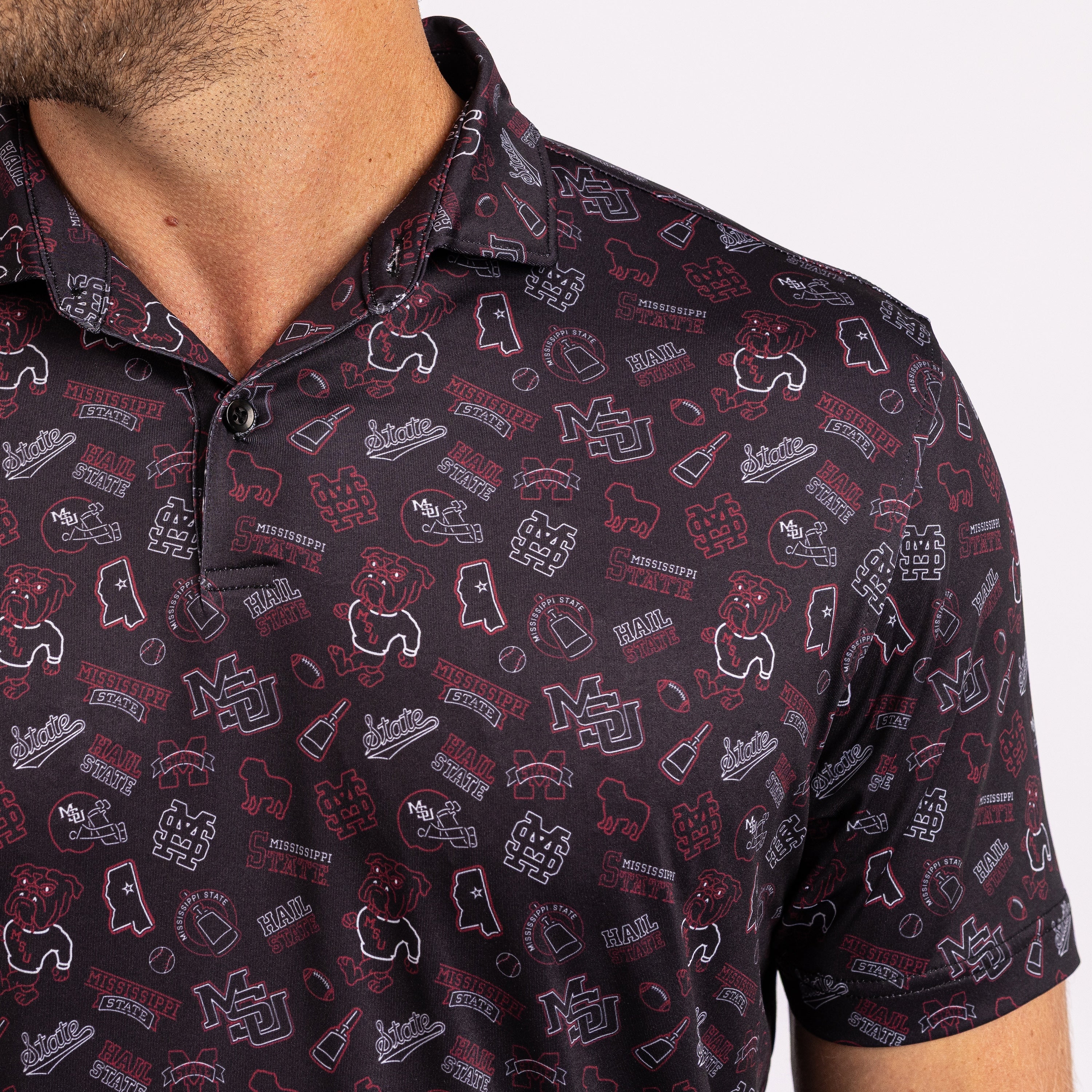 A close-up of a man wearing a short-sleeve polo shirt with an all-over Mississippi State Bulldogs collegiate graphic print. The shirt features a dark navy base with red and white logos, including the bulldog mascot
