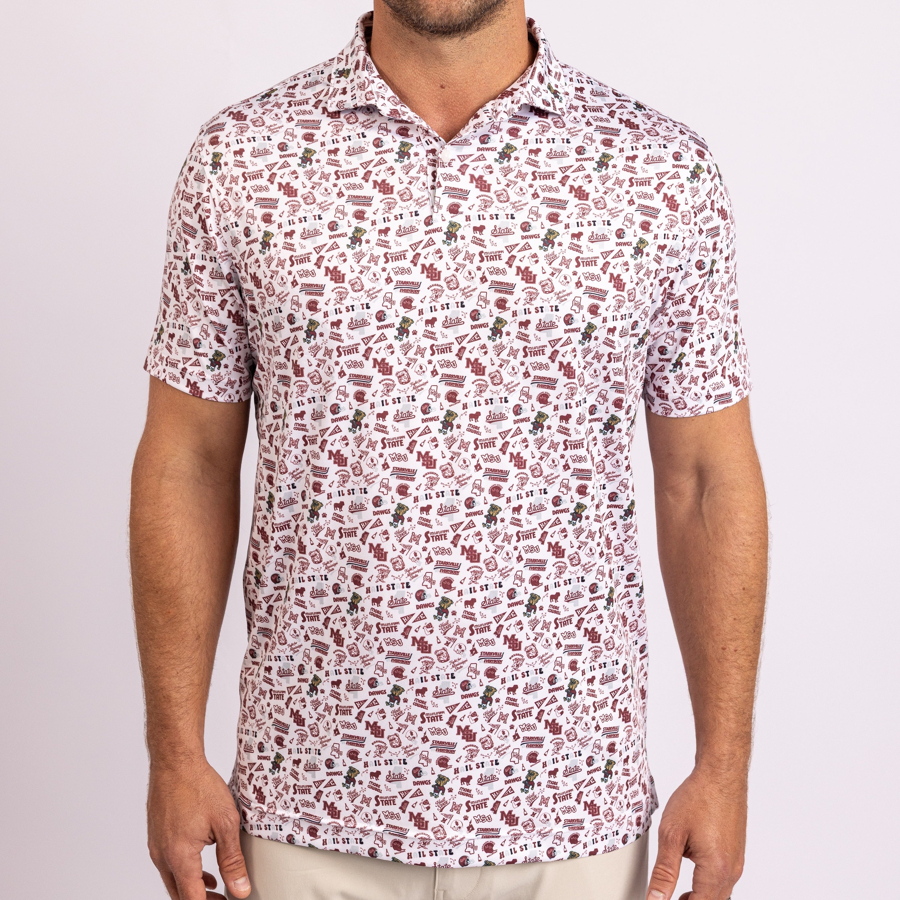A man wearing a short-sleeve polo shirt with an all-over abstract pattern of red and brown shapes on a white background, paired with light beige pants, against a light gray studio background.