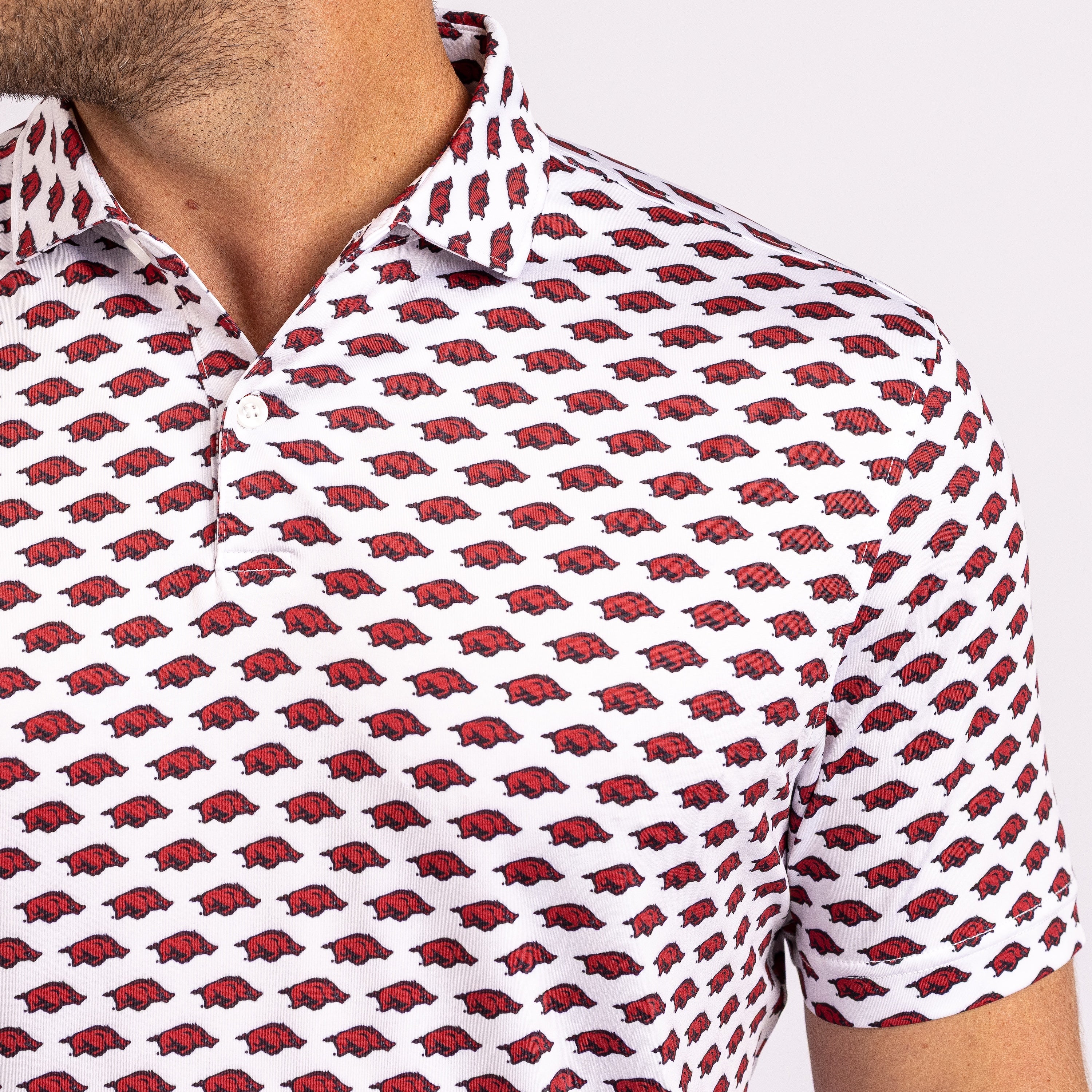 A close-up of a man wearing a white short-sleeve polo shirt with an all-over repeating red and black Arkansas Razorbacks logo pattern.