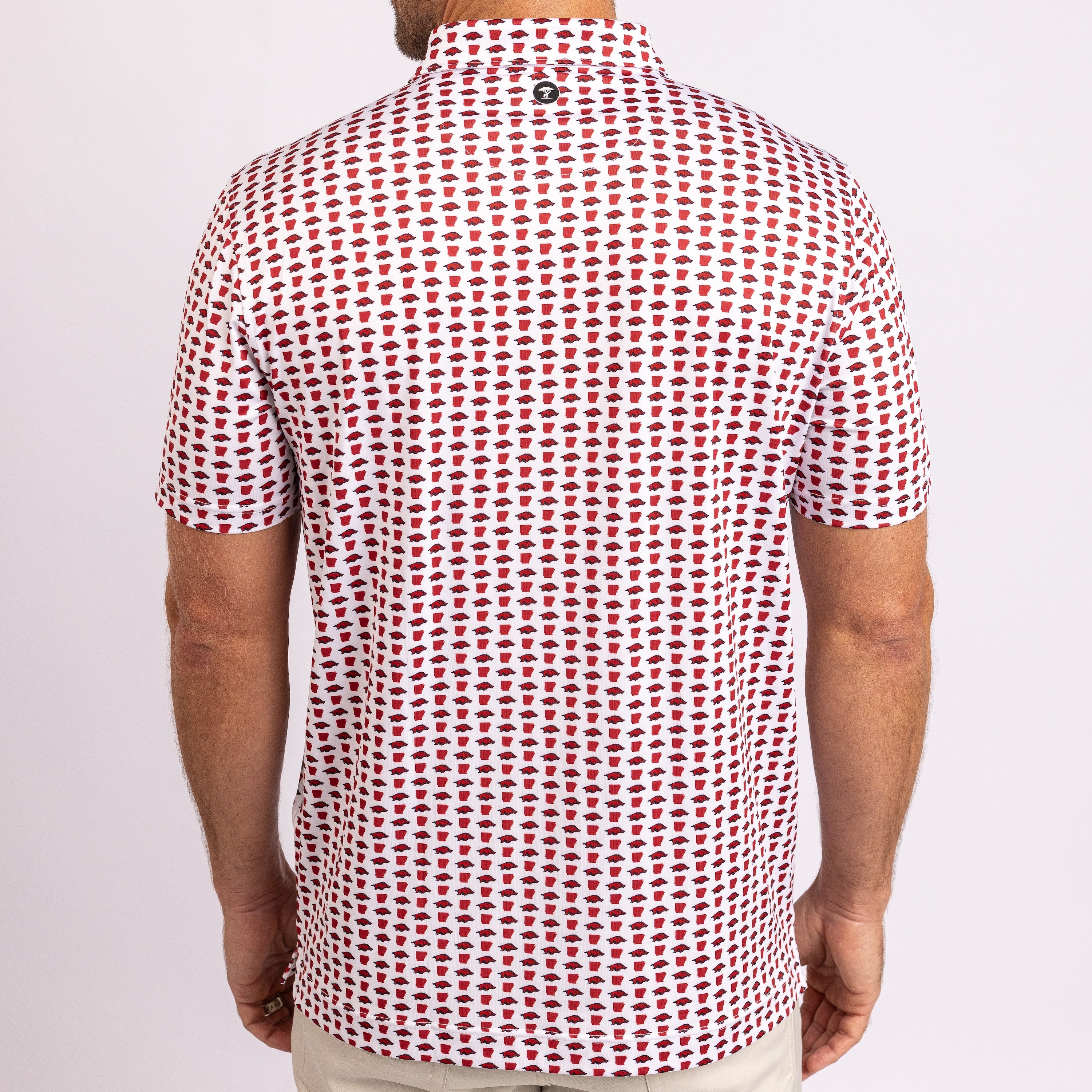 A man viewed from the back wearing a short-sleeve polo shirt with a repeating red and black geometric pattern on a white base, paired with light beige pants against a light gray studio background.