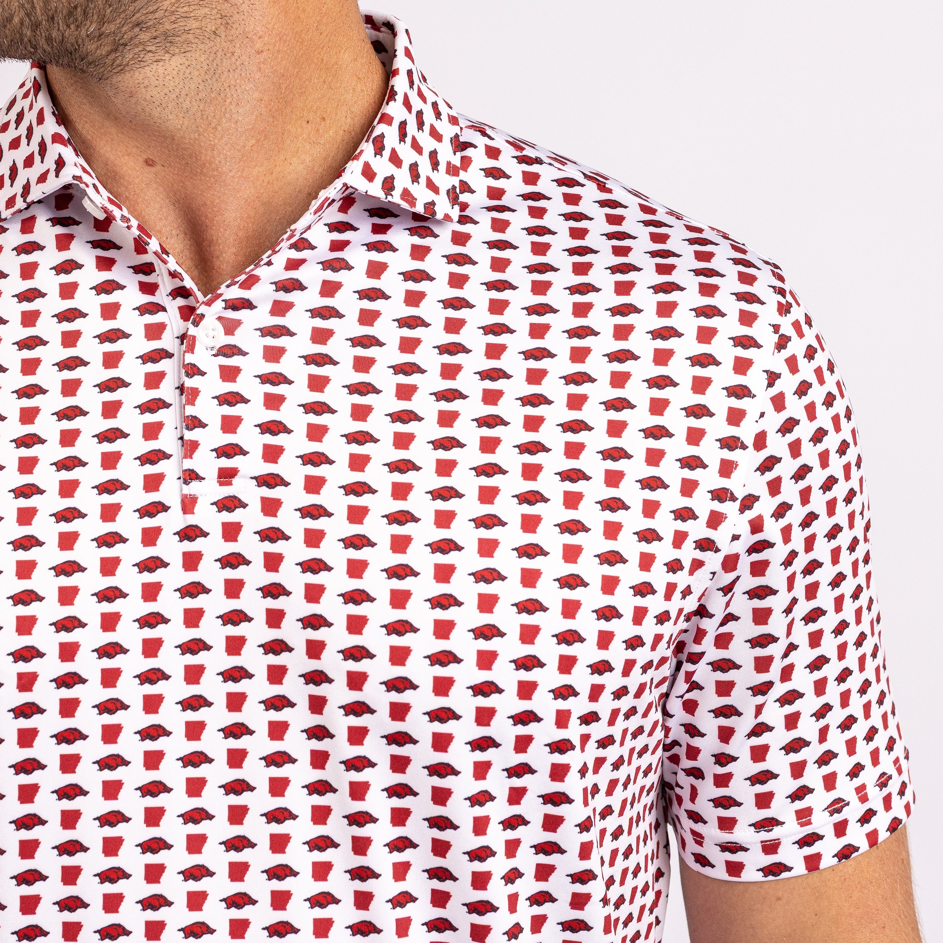 A close-up of a man wearing a white short-sleeve polo shirt with an all-over repeating pattern of red and black Arkansas Razorbacks logos.