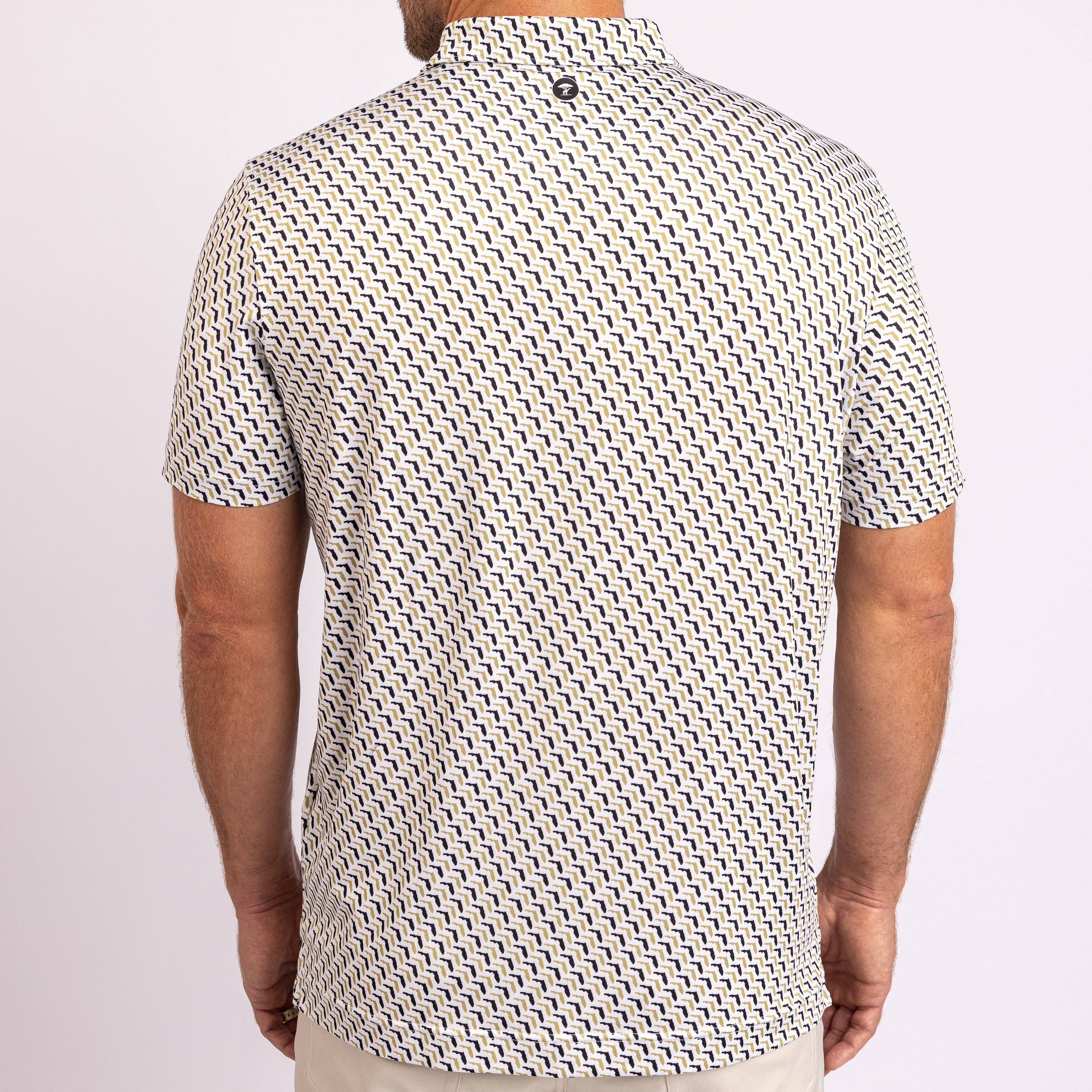 A man viewed from the back wearing a short-sleeve button-up shirt with a repeating black and white geometric pattern, paired with light beige pants, against a plain light background.