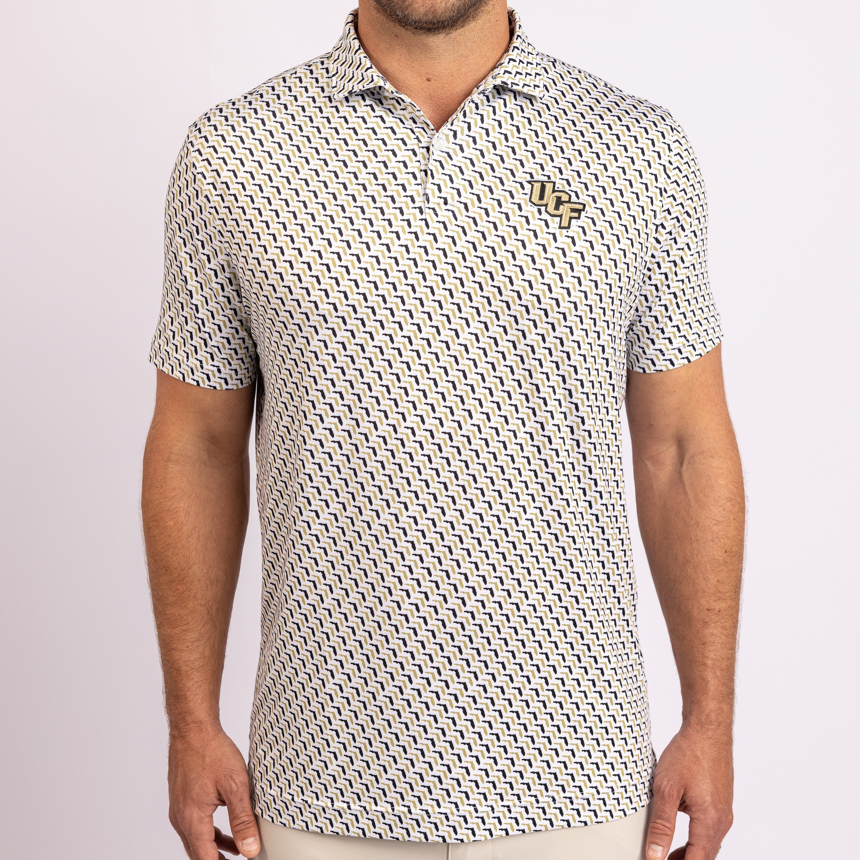 A man wearing a short-sleeve polo shirt with a repeating black and white geometric pattern and a small gold embroidered logo on the left chest, photographed against a light gray studio background.