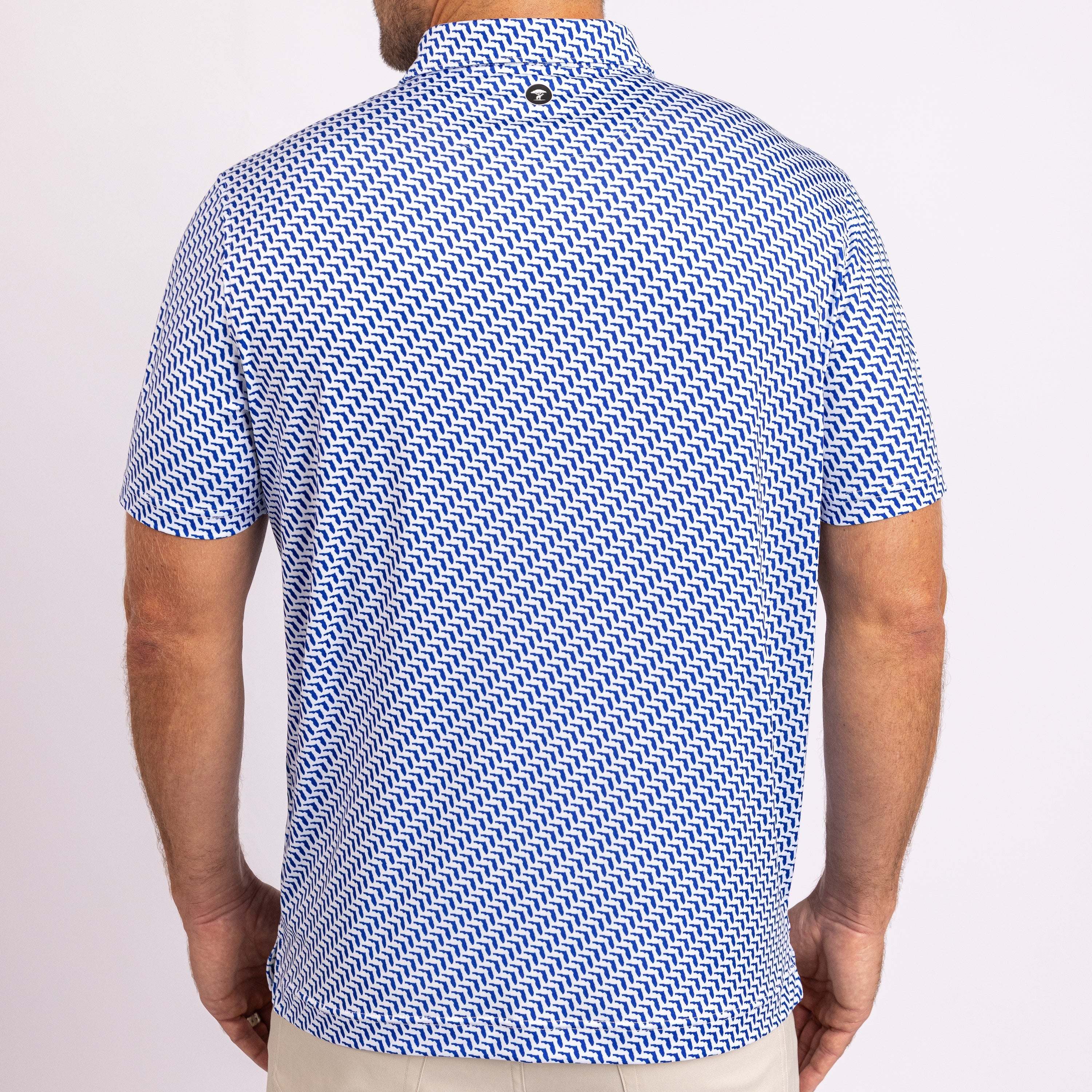 A man viewed from the back wearing a short-sleeve polo shirt with a blue and white geometric zigzag pattern, paired with beige pants, against a light studio background.