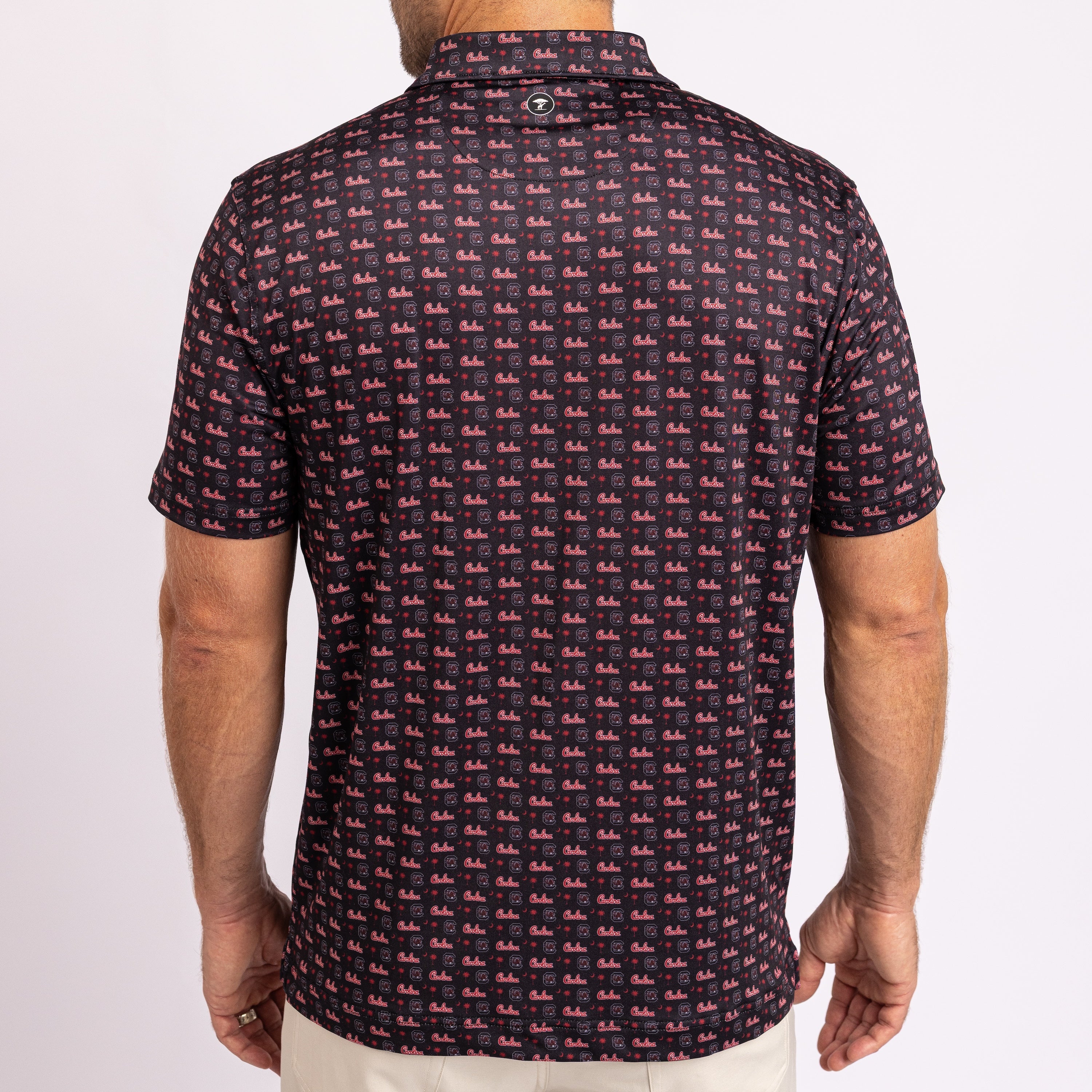 A man viewed from the back wearing a short-sleeve collared polo shirt with an all-over repeating pink and dark navy pattern, paired with light beige pants against a plain light background.