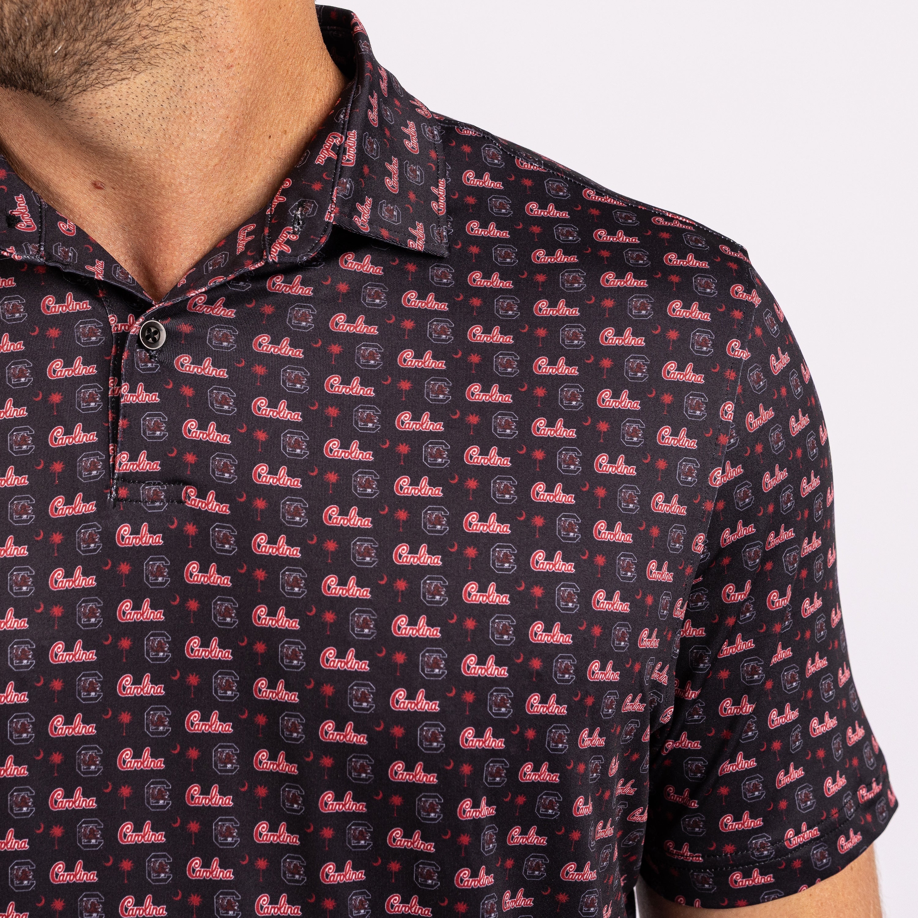 Close-up of a man wearing a short-sleeve polo shirt with an all-over repeating 'Coach' logo print in red and white on a dark navy background.