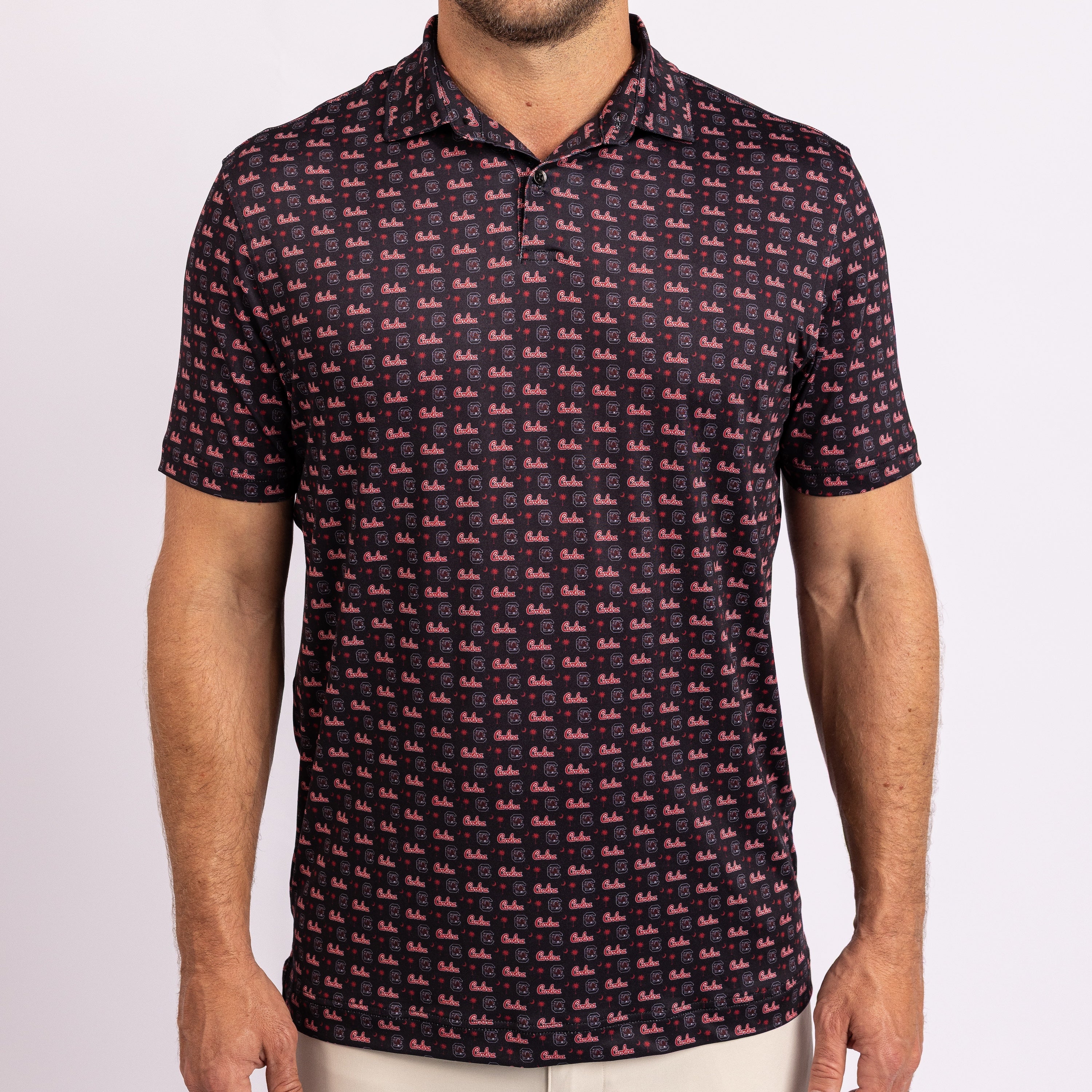 A man wearing a short-sleeve polo shirt with an all-over repeating pink and red logo pattern on a dark navy background, paired with light beige pants, against a plain light gray studio background.