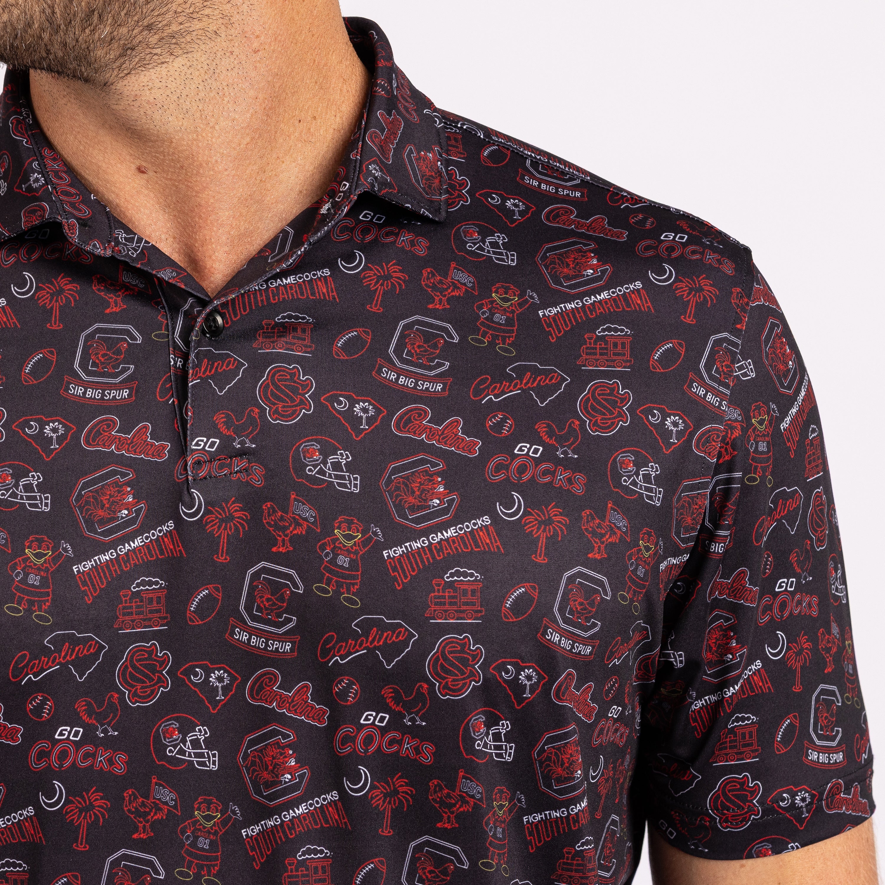 A close-up of a man wearing a short-sleeve polo shirt with an all-over collegiate graphic print. The shirt features red, white, and gray football-themed illustrations and text, including South Carolina Gamecocks