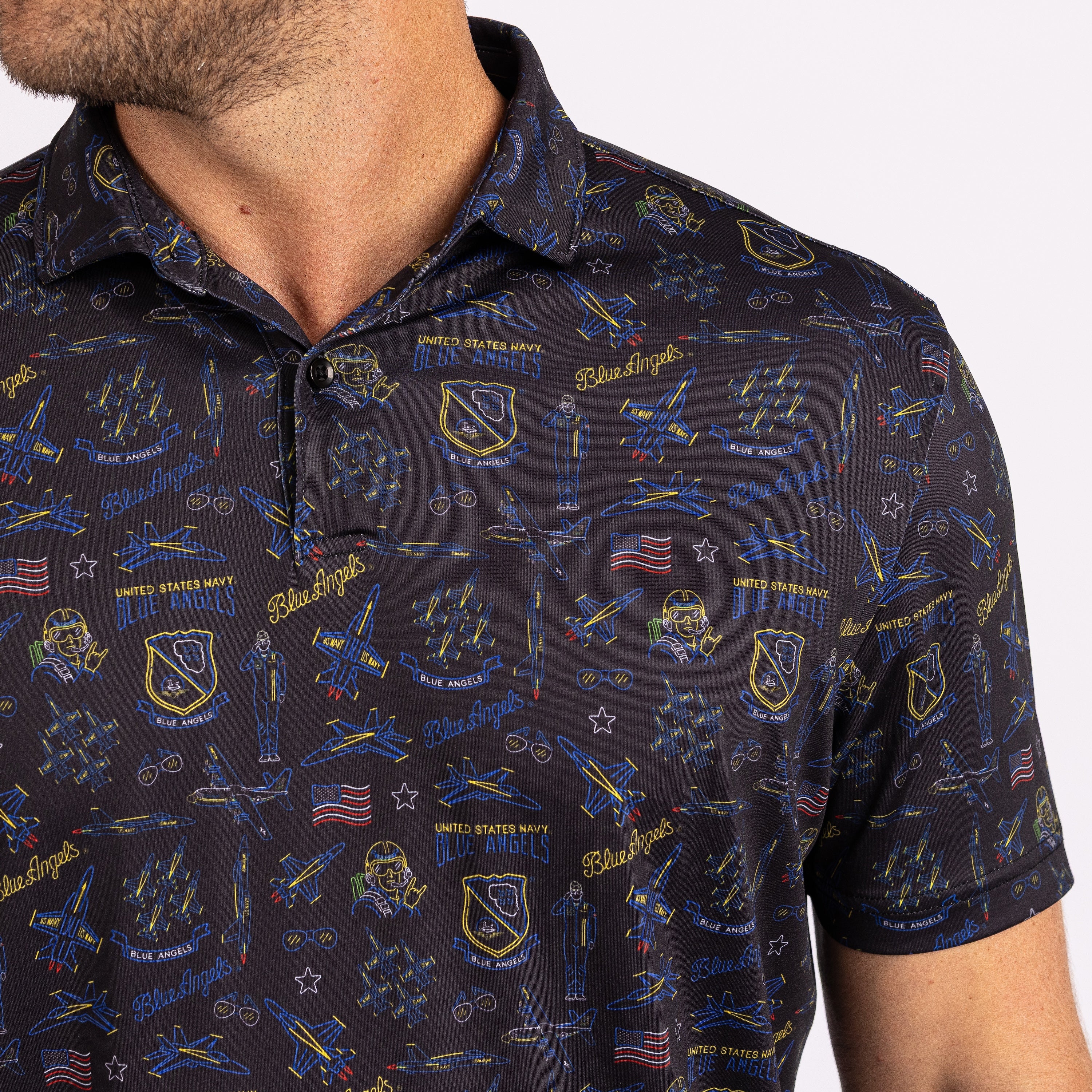 A close-up of a man wearing a short-sleeve polo shirt with a dark navy blue base and an all-over United States Navy Blue Angels themed print featuring blue, gold, and red details.
