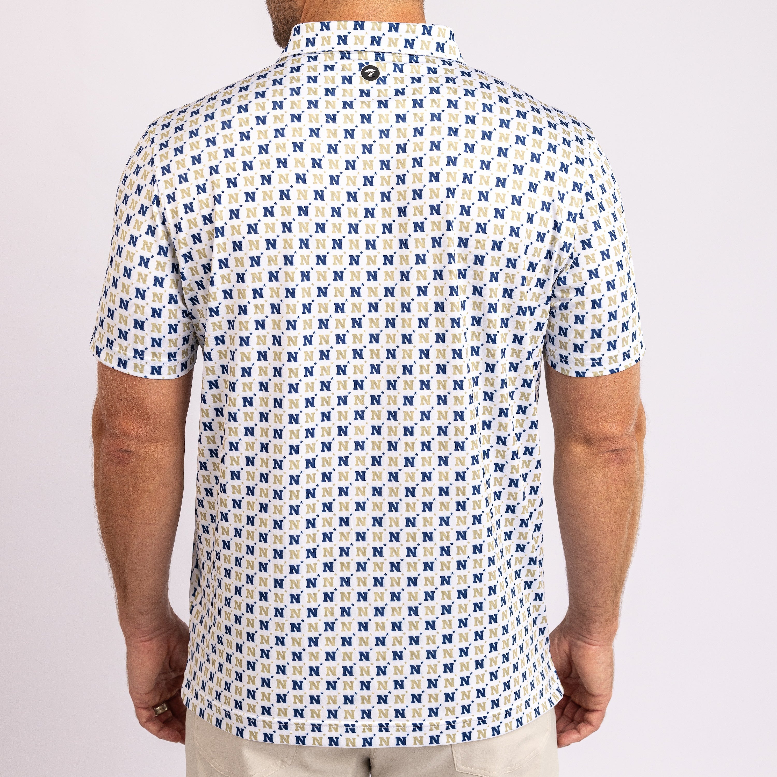 A man viewed from the back wearing a short-sleeve button-up shirt with an all-over repeating blue and tan monogram pattern on a white base, paired with light beige pants against a light gray studio background.