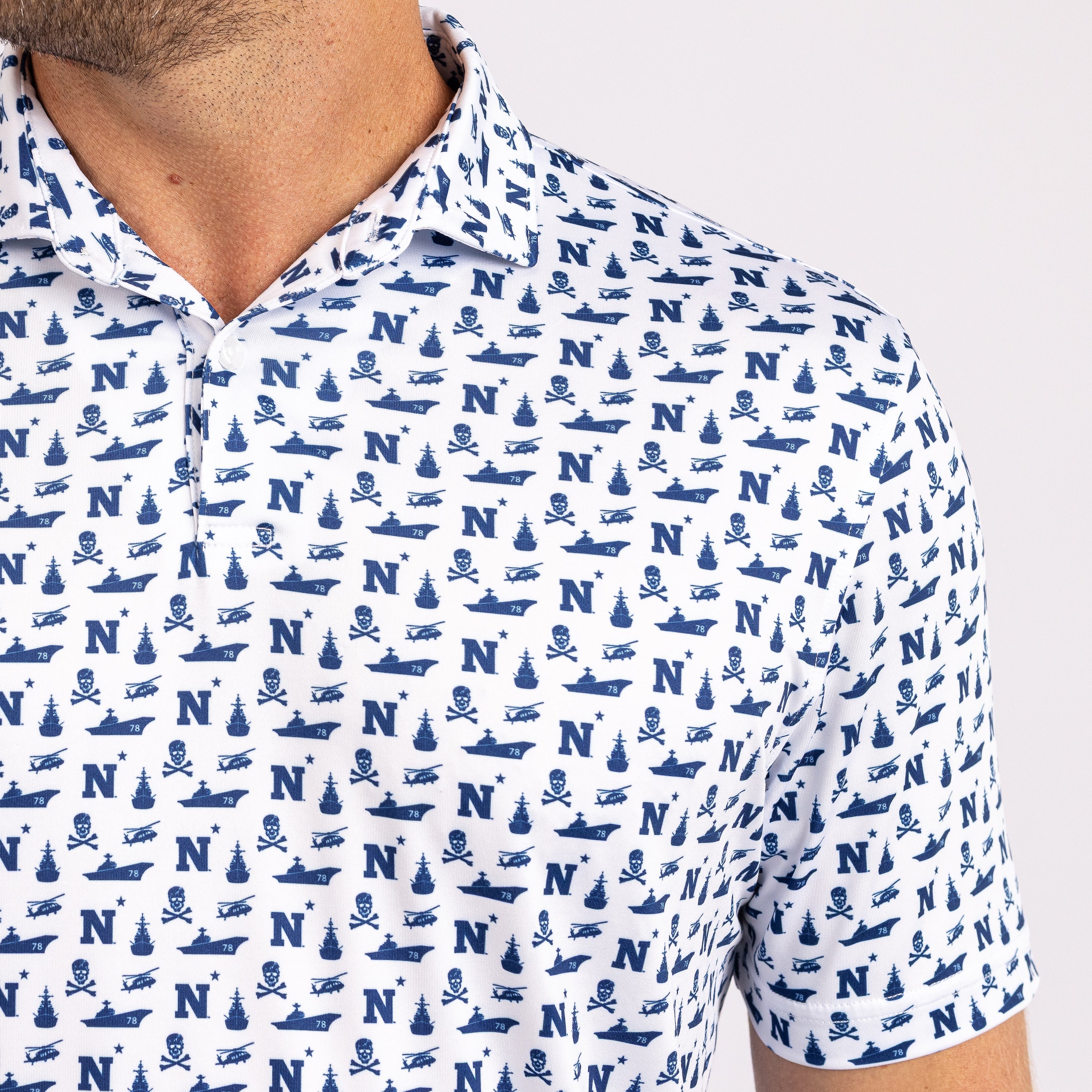 A close-up of a man wearing a white short-sleeve polo shirt with an all-over navy blue pattern of the letter 'N' and nautical icons, set against a plain light gray background.