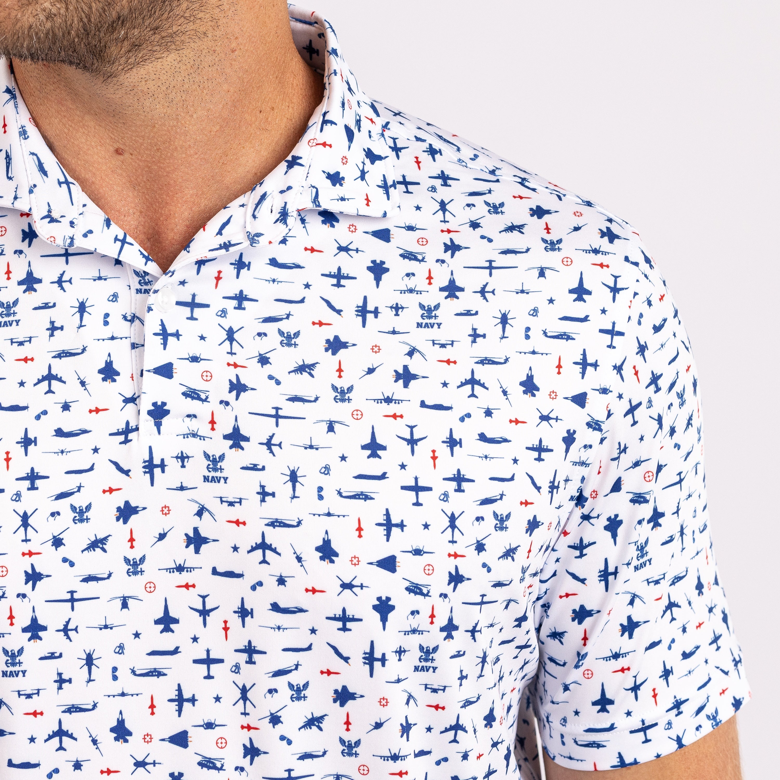 A close-up of a man wearing a white short-sleeve polo shirt with an all-over pattern of blue and red military aircraft and ships.
