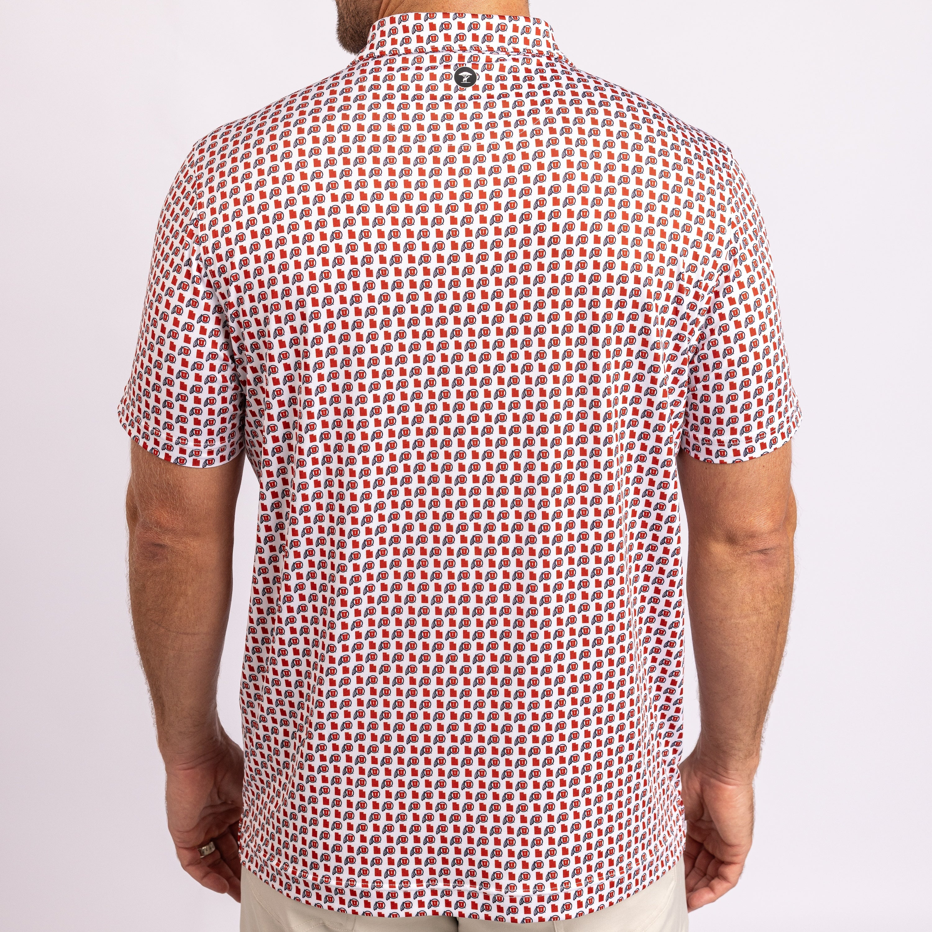 A man viewed from the back wearing a short-sleeve button-up shirt with a dense repeating geometric pattern in red, white, and black, paired with light beige pants against a plain light background.