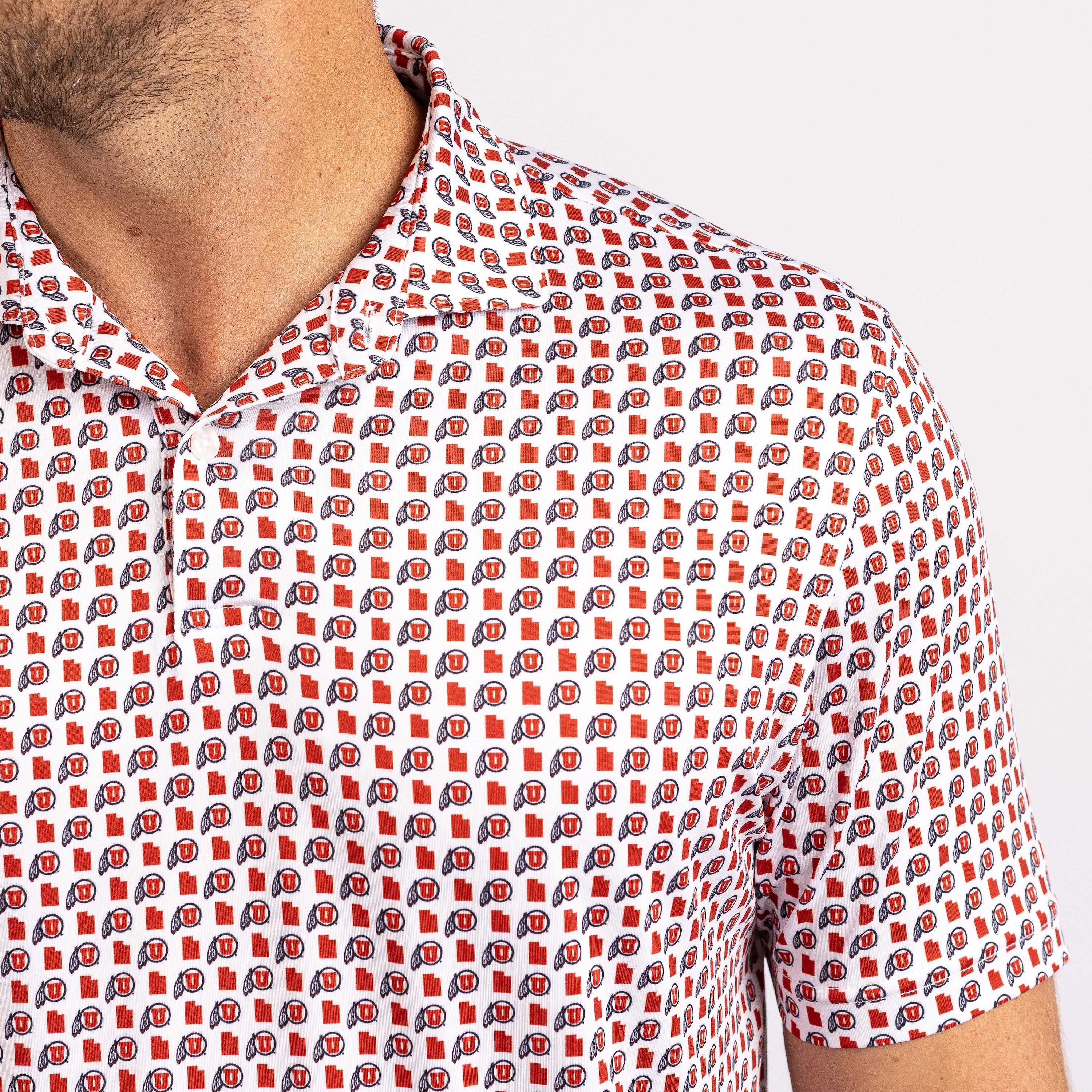 Close-up of a man wearing a short-sleeve collared polo shirt with an all-over repeating red, navy, and white logo pattern on a white base.