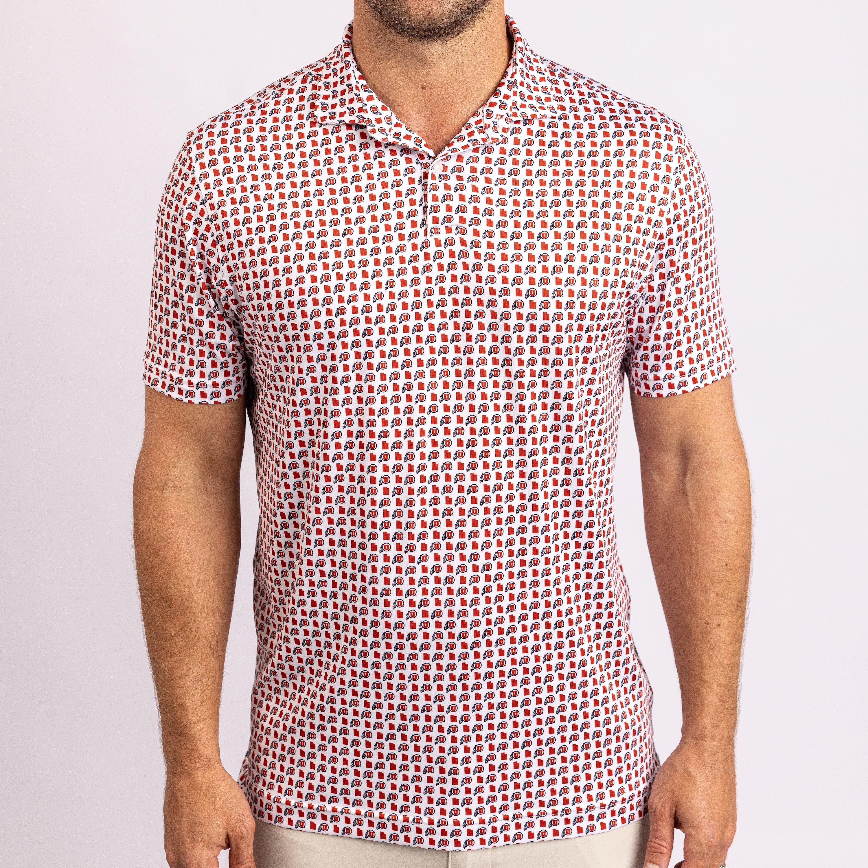 A man wearing a short-sleeve polo shirt with a dense, repeating geometric pattern of red, white, and black shapes on a light base, paired with light beige pants against a plain light background.