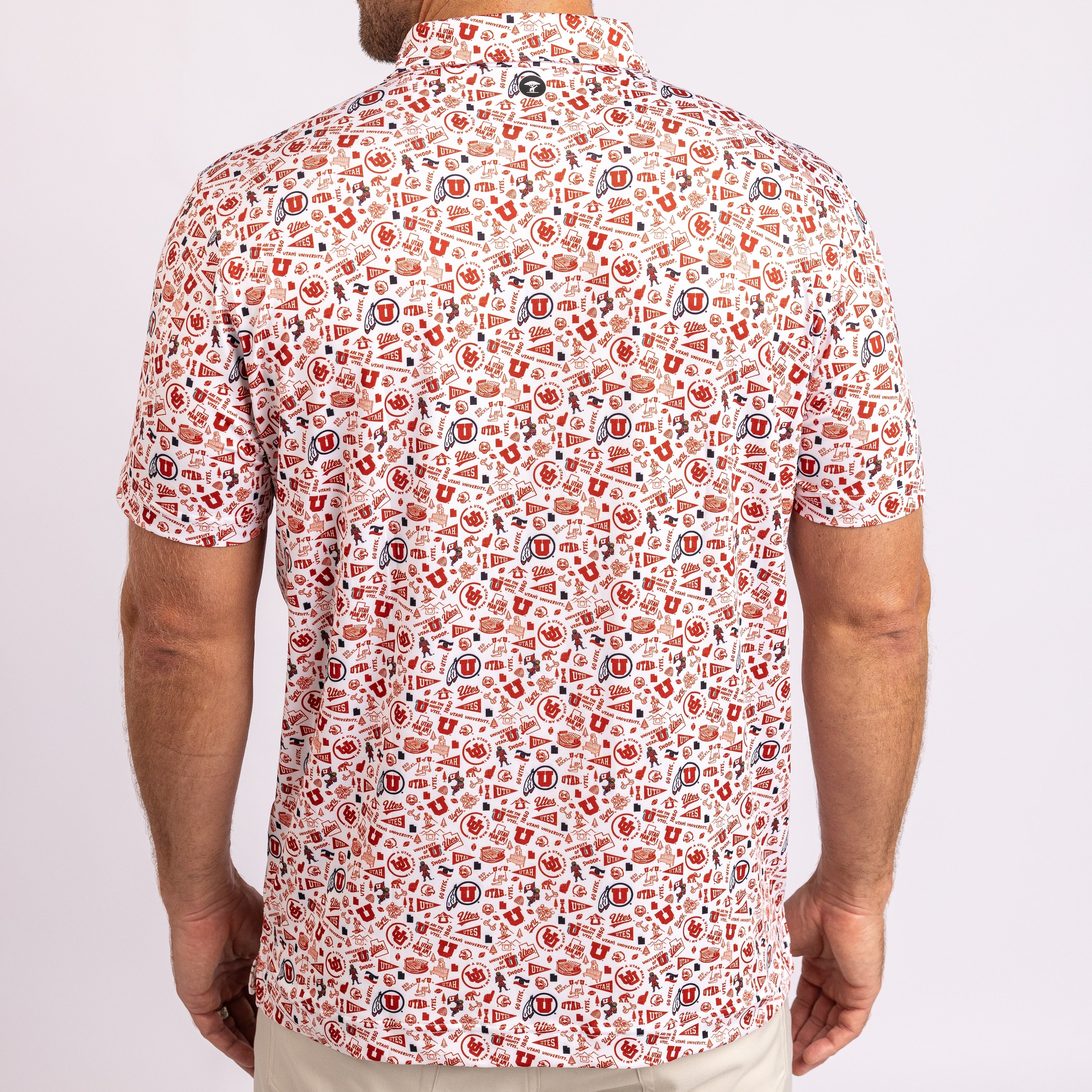 A man viewed from the back wearing a short-sleeve button-up shirt with an all-over collegiate-style graphic print in red, blue, and black on a white base, paired with light khaki pants against a light gray studio background.