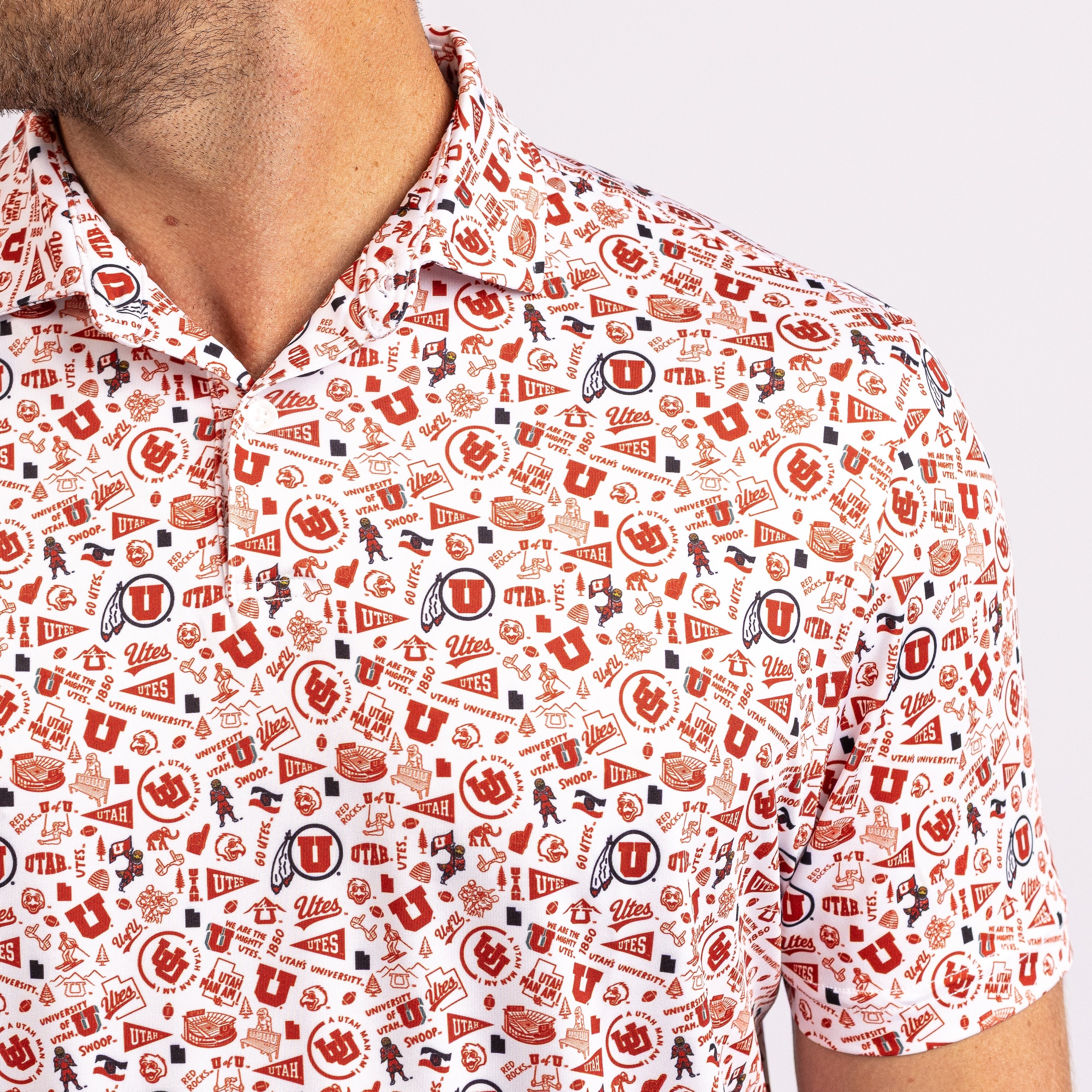 A close-up of a man wearing a short-sleeve polo shirt with an all-over collegiate-style print in red, navy, and black on a white base. The print features letters, logos, flags, and sports icons, including a large 'U' text.