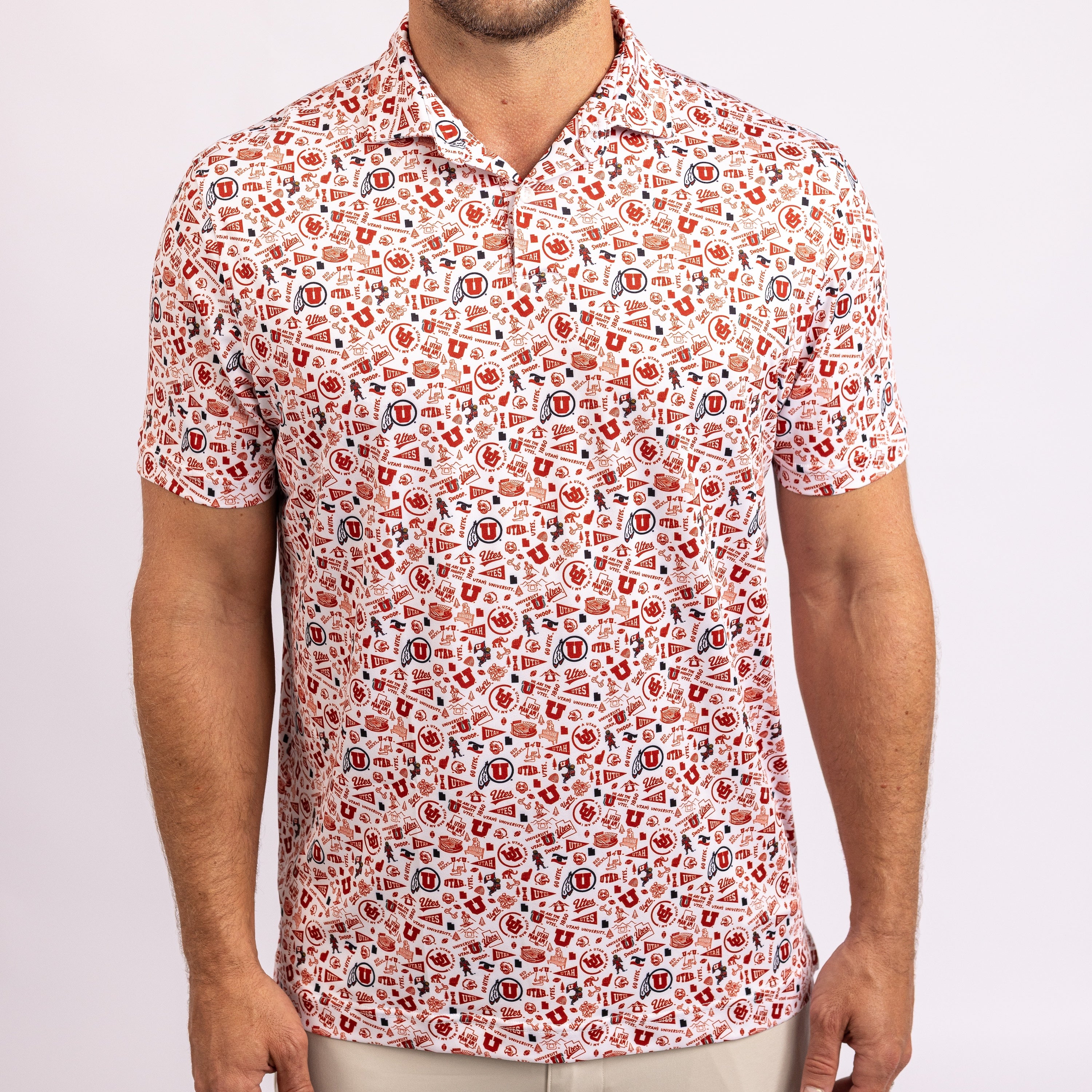 A man wearing a short-sleeve polo shirt with an all-over collegiate-style pattern of red, blue, and black lettering and logos on a white background, photographed against a light gray studio backdrop.