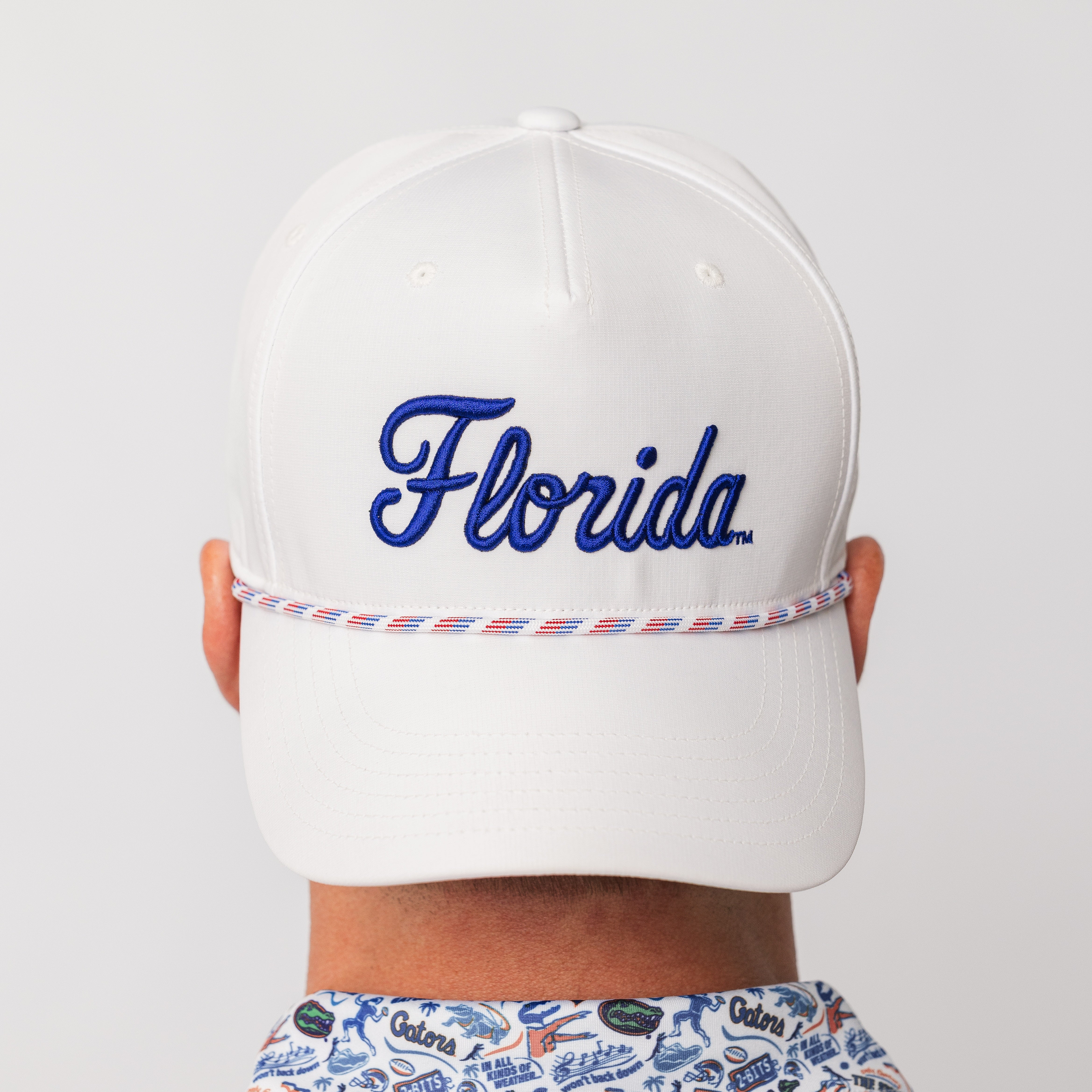 A white baseball cap with 'Florida' embroidered in blue script on the front, shown from the back on a model. The cap features a thin red and white rope detail around the crown and a patterned shirt underneath.
