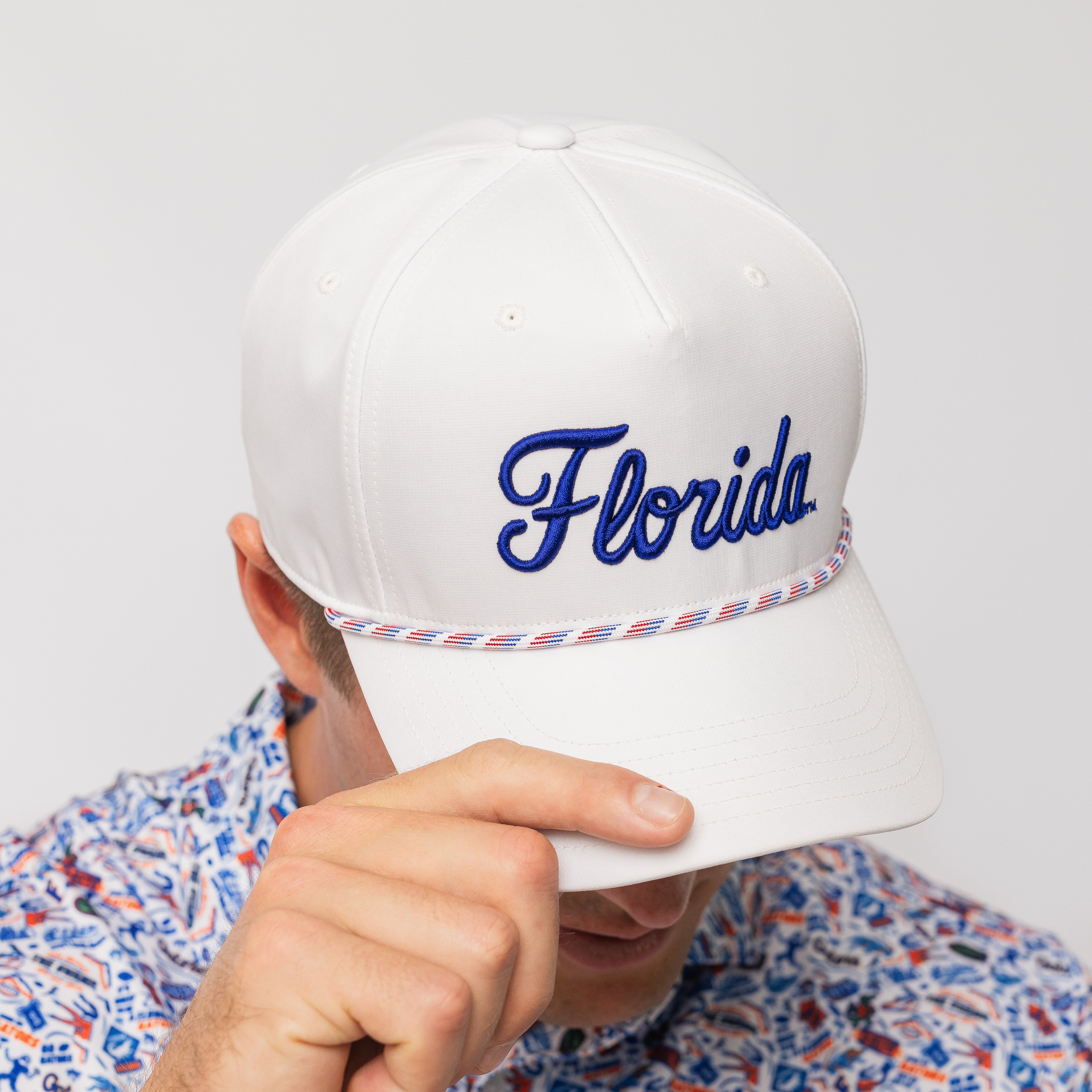 A white baseball cap with 'Florida' embroidered in blue script on the front, featuring a red, white, and blue braided rope detail around the brim. A person is wearing it and holding the brim down, with a patterned shirt visible.